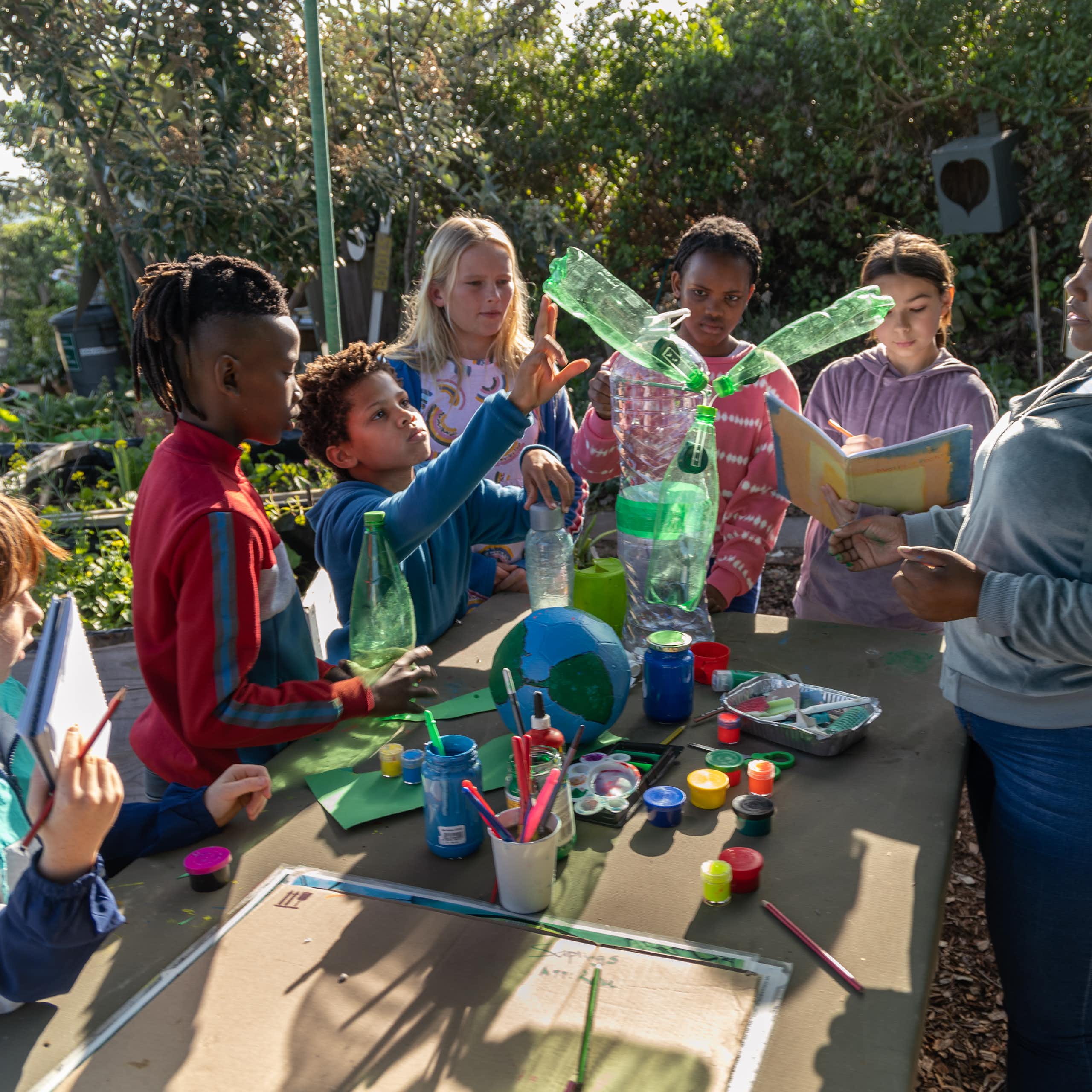 Six young children in casual clothes, some holding books and others paintbrushes, gather around a model of the earth and some plastic bottles fitted together while an adult, their teacher, talks to them