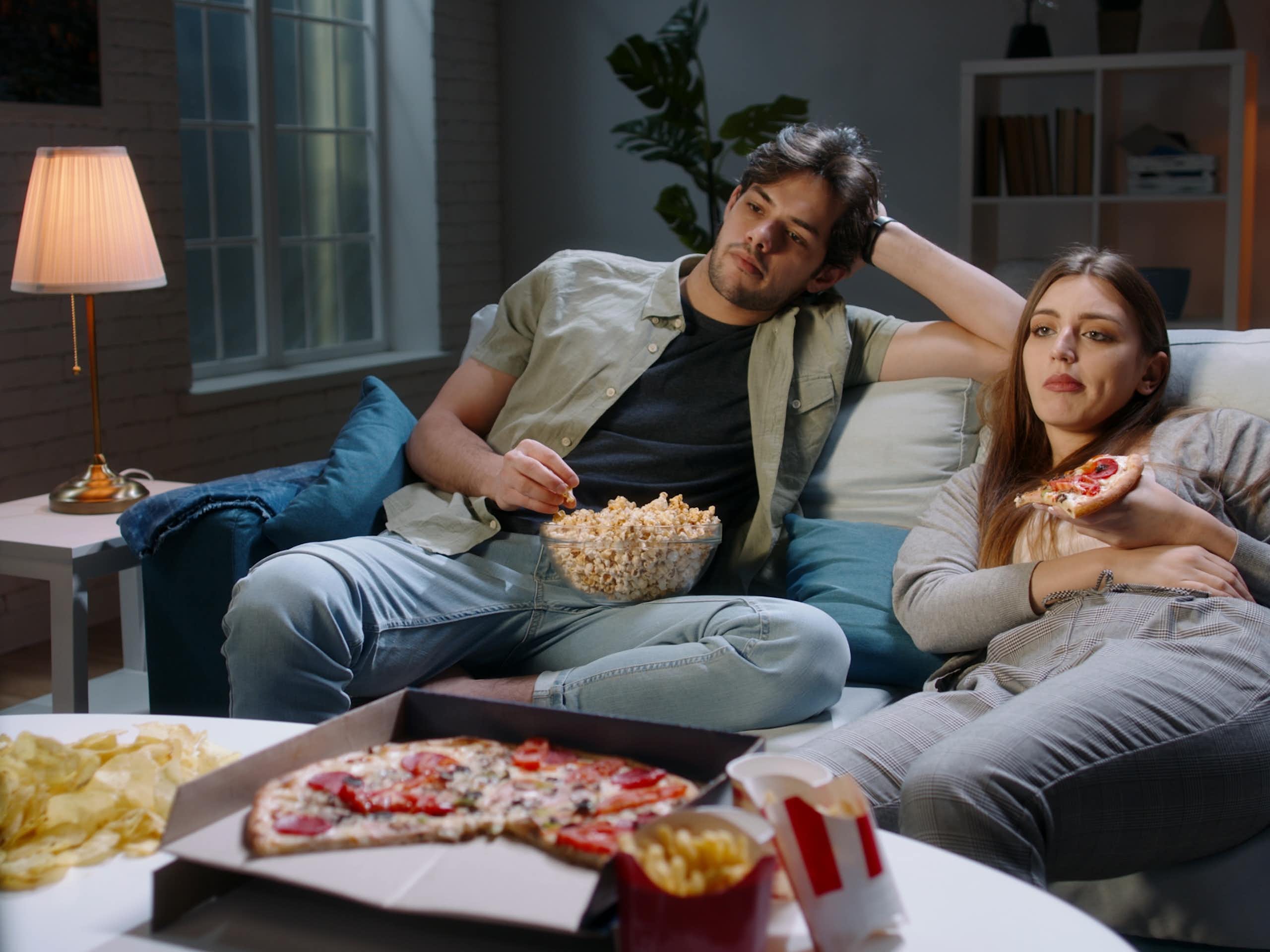 Un couple est assis sur un sofa, en train de manger, l'air ennuyé