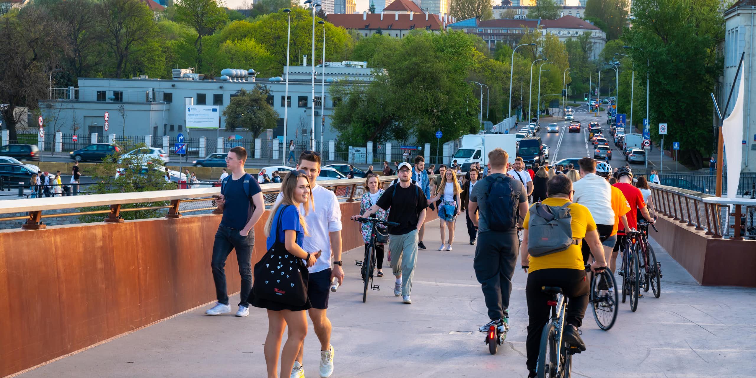 A group of people, some on bicycles, cross a bridge, with skyscrapers in the background.
