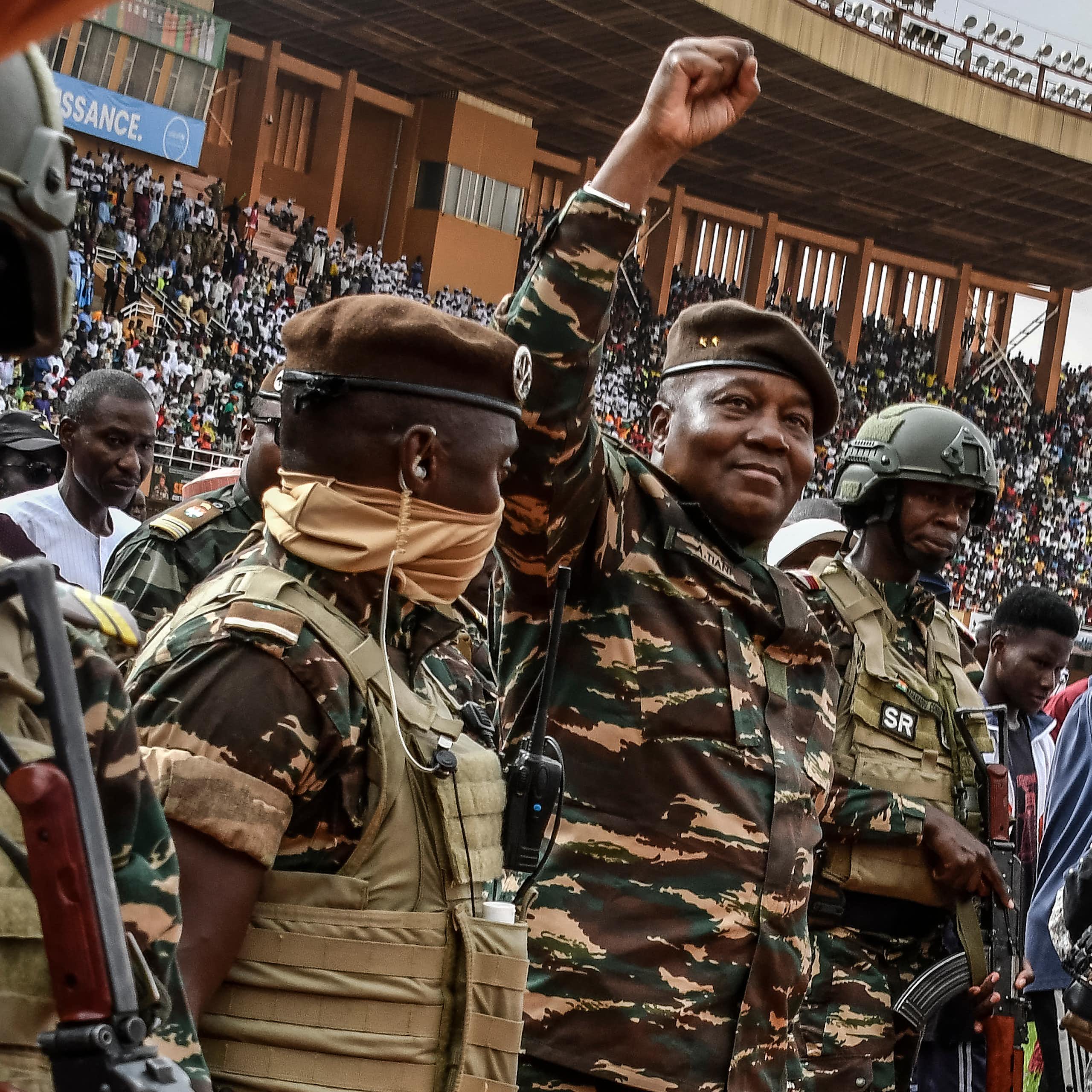 A smiling man dressed in army fatigues holding up a fisted arm while surrounded by armed military men, with a crowd of seated people behind him in a stadium