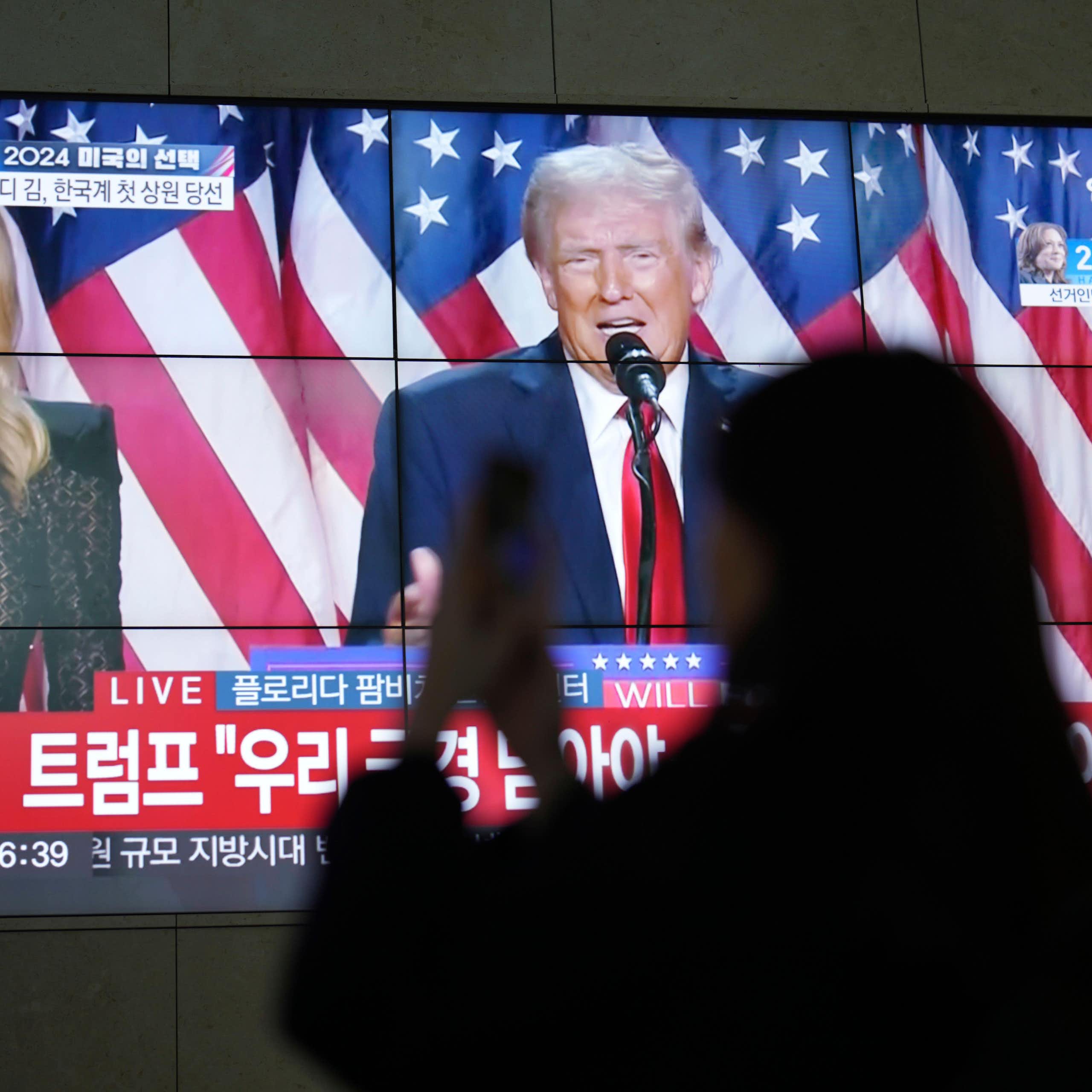 A silhouette of a woman looking at her phone with Donald Trump on a TV screen behind her.