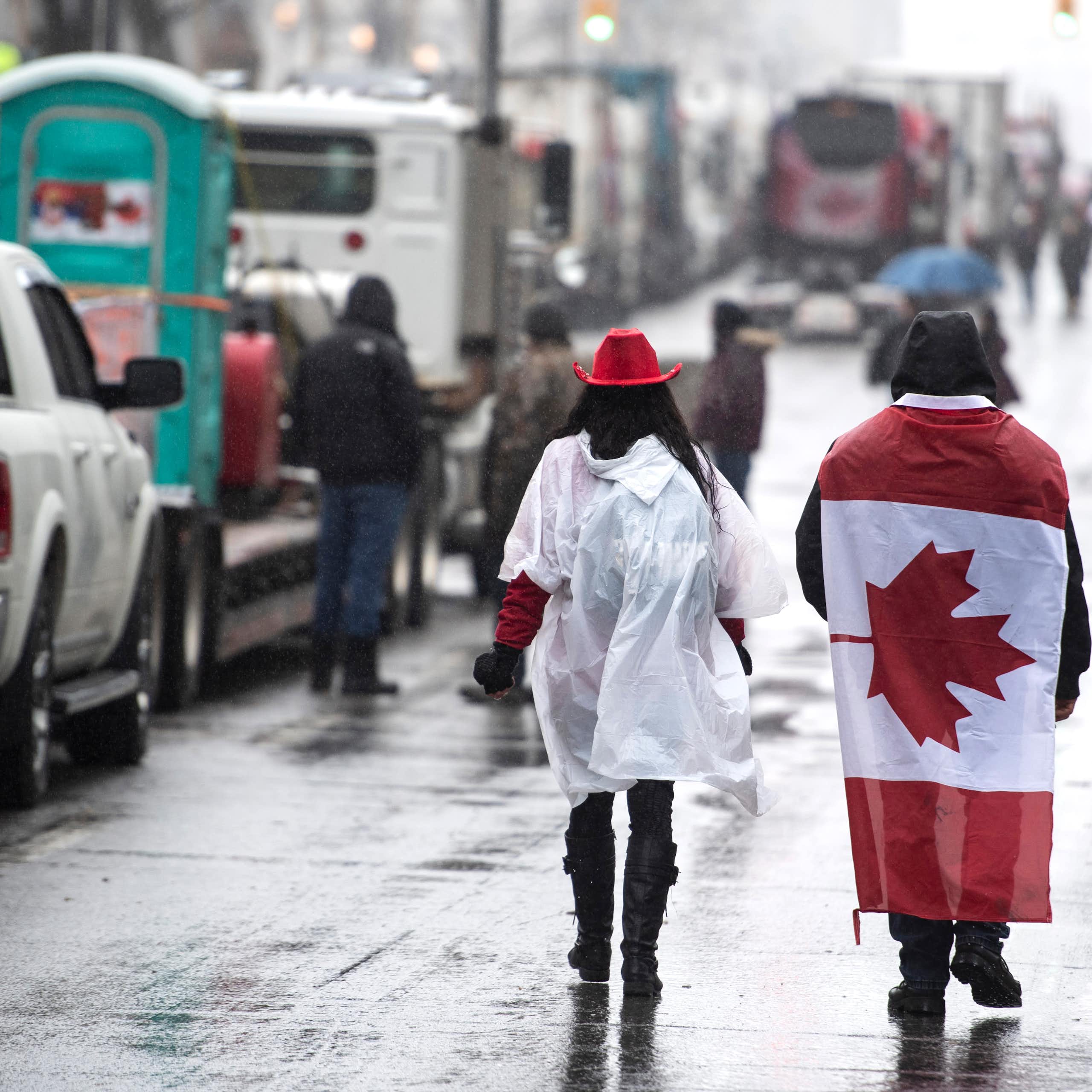 People walk down a rainy street lined with cars, one of them wears a canadian flag as a cape 