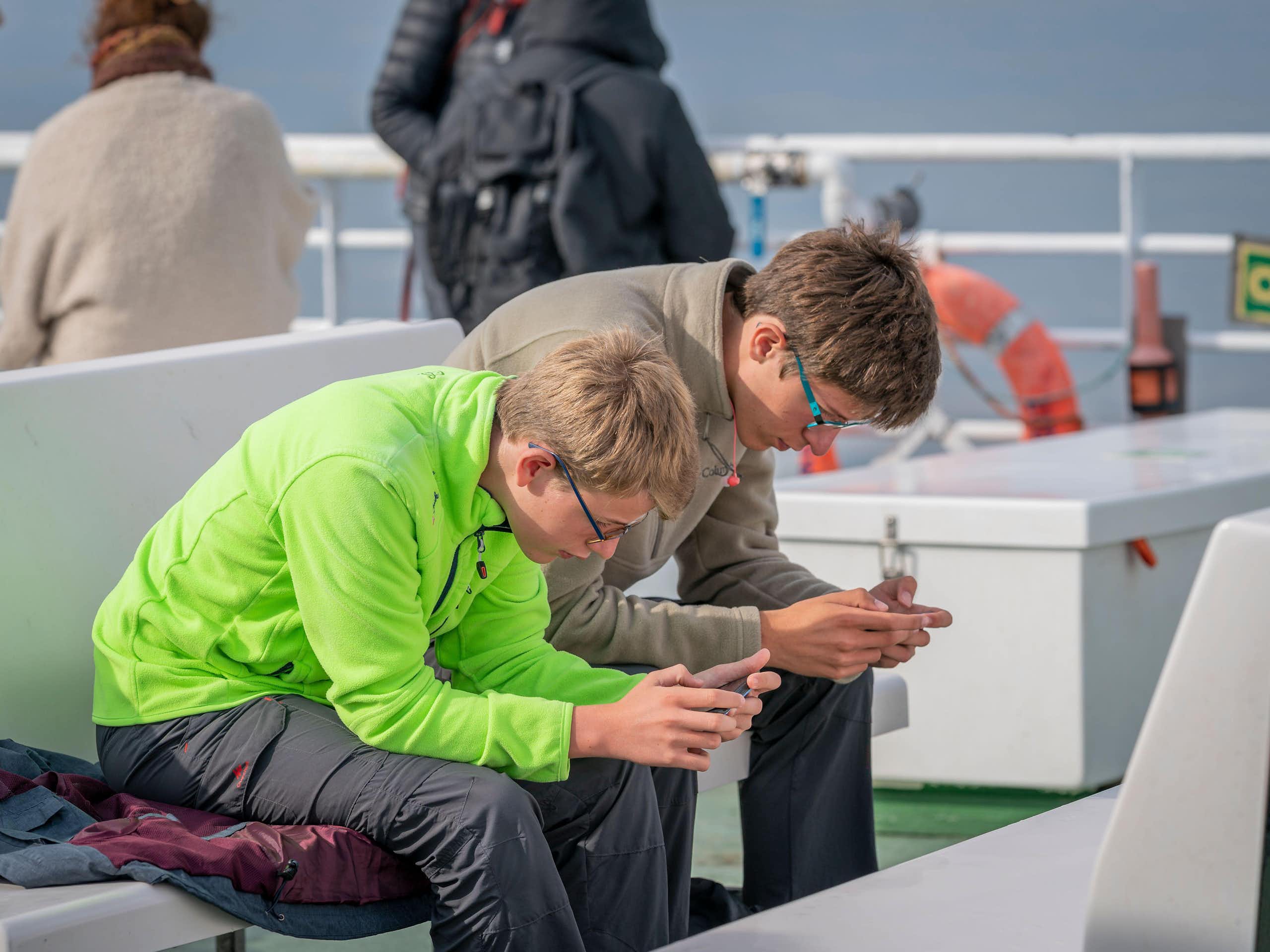 Two teenager boys on a ferry bent over, staring at their phones.