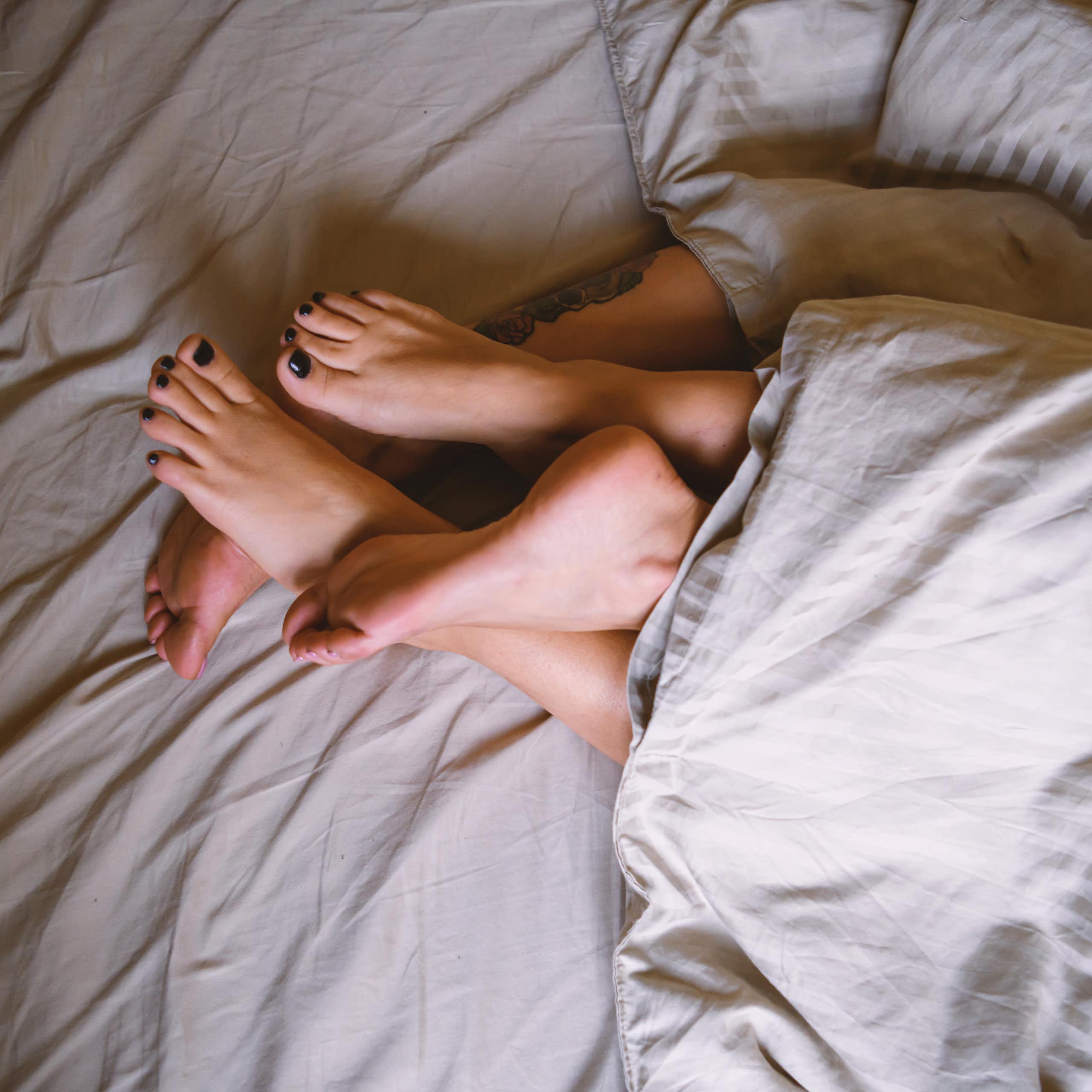 Intertangled feet of two women peek out from a duvet cover.