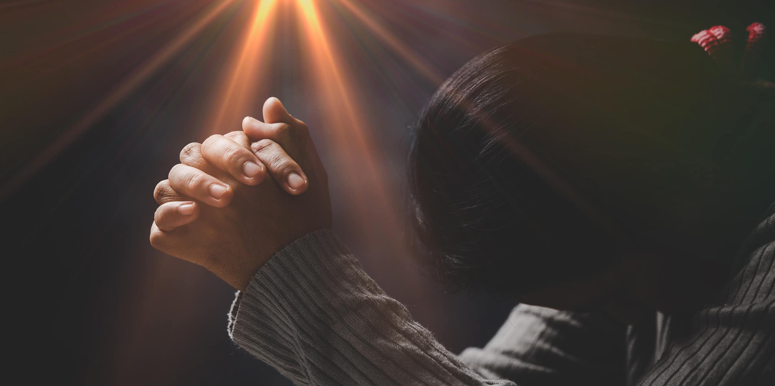 A woman with folded hands against a black background with a yellow light coming from above them