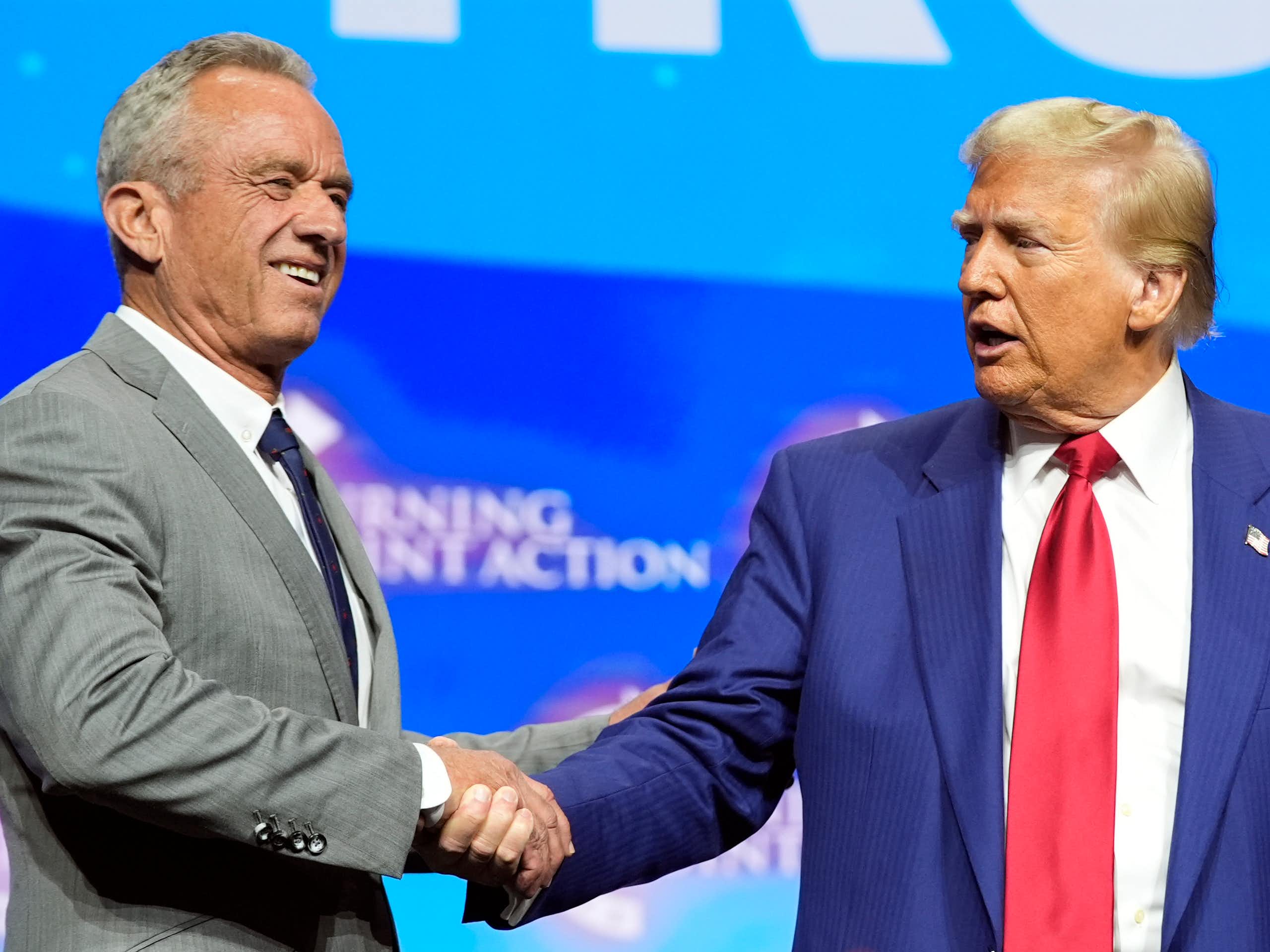 A man in a light gray suit shakes hands with a man in a navy suit and red tie