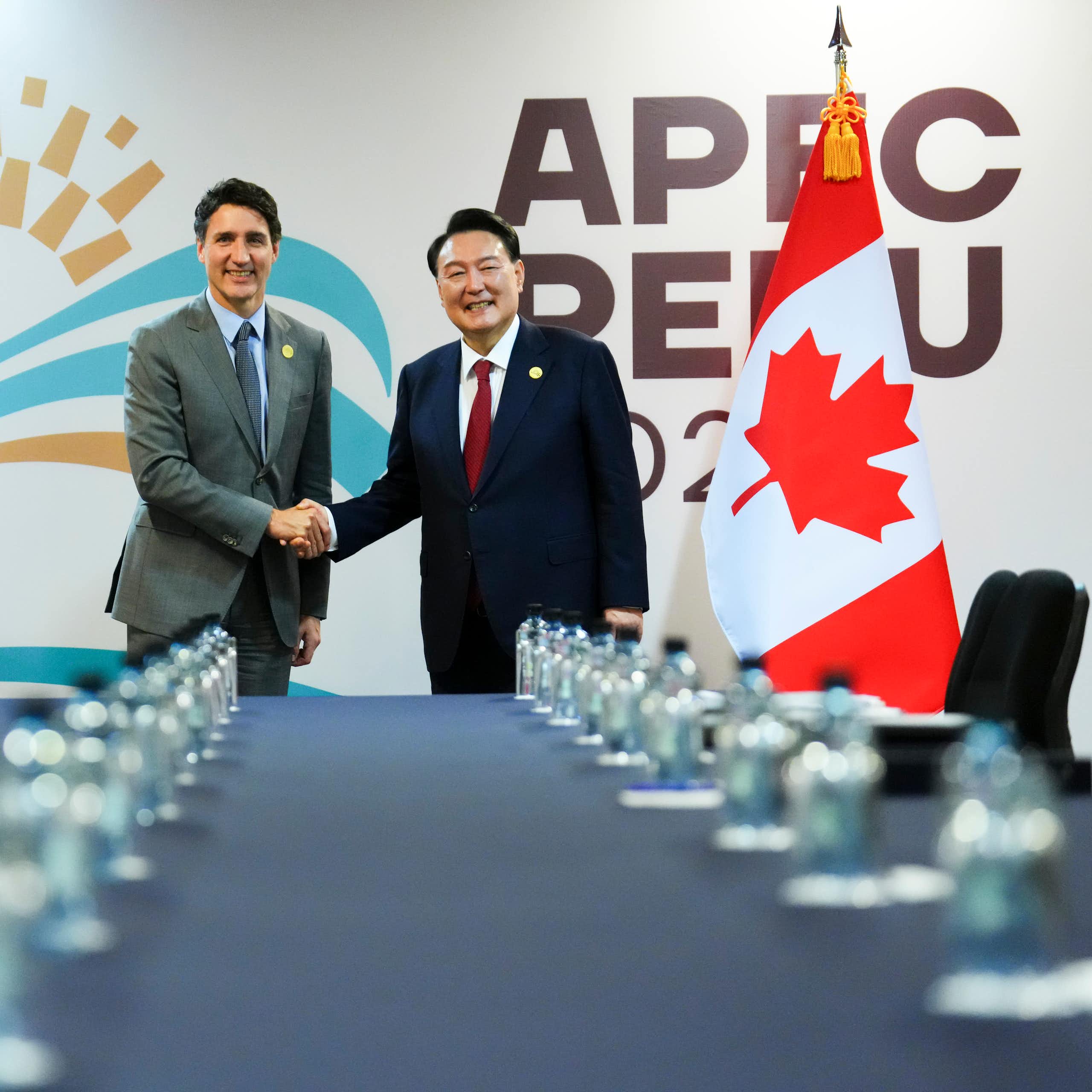Two smiling dark-haired men, one white and one Asian, shake hands at the end of a table lined with water bottles.