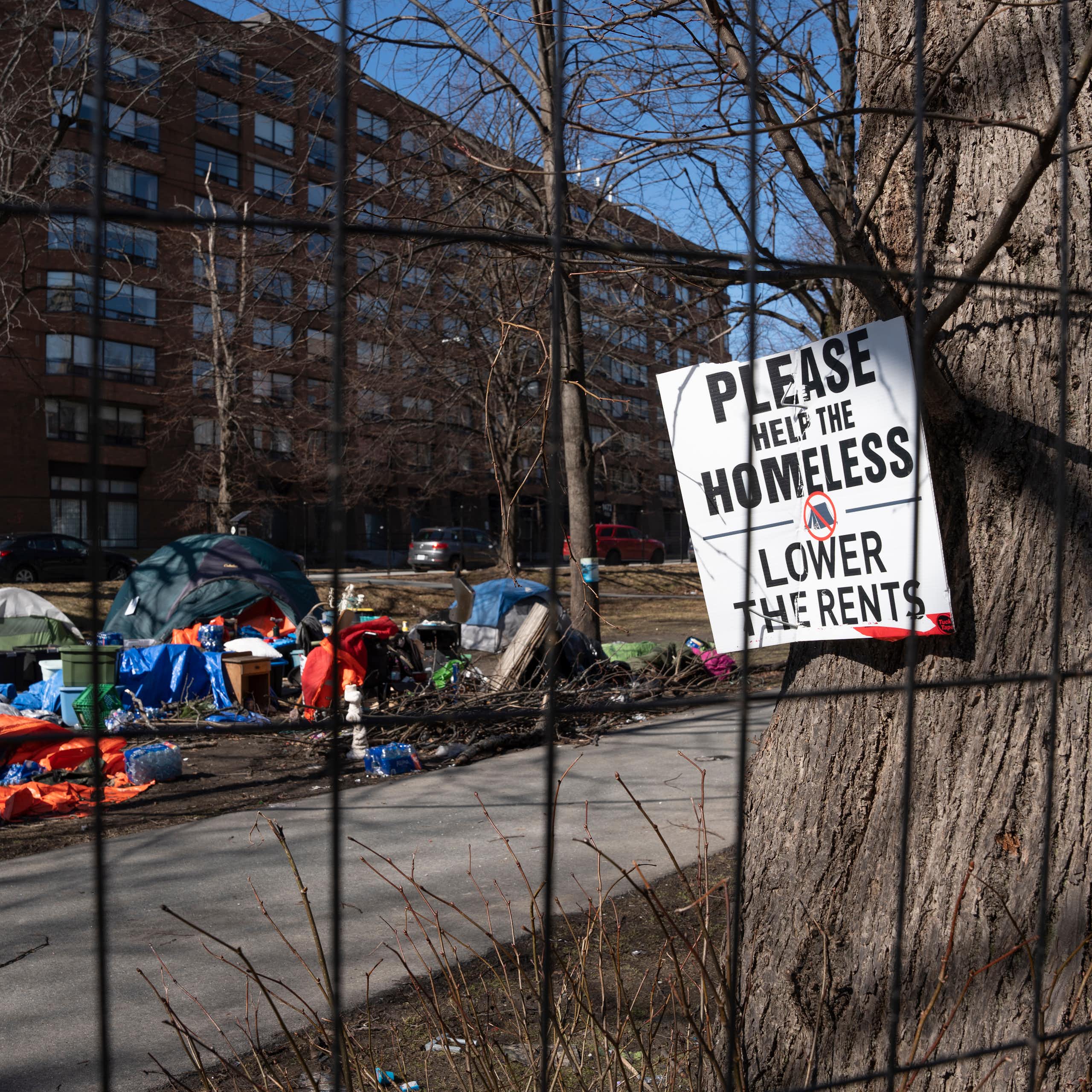 A sign pinned to a tree near a homeless encampment reads Please Help The Homeless, Lower the Rents