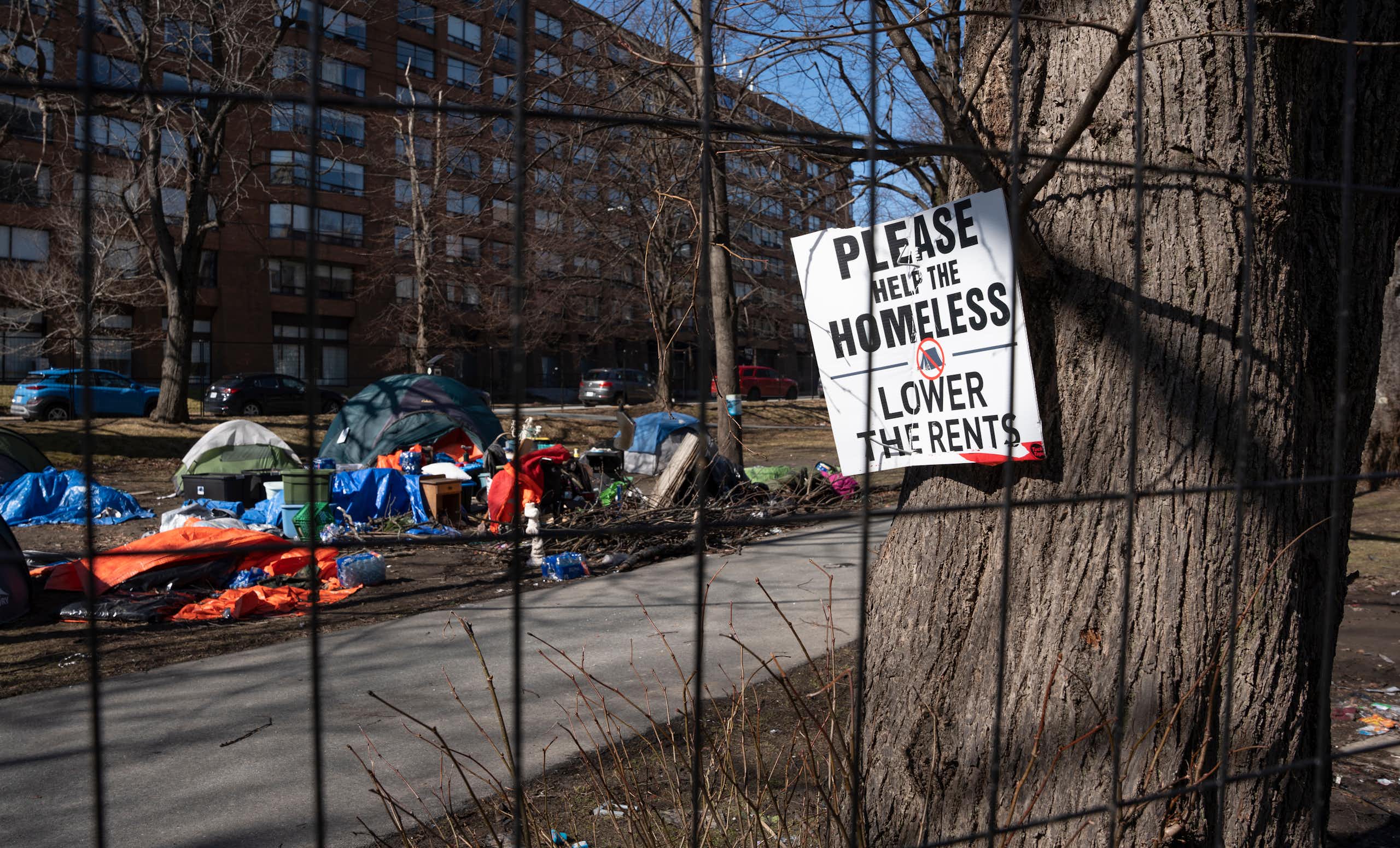 A sign pinned to a tree near a homeless encampment reads Please Help The Homeless, Lower the Rents