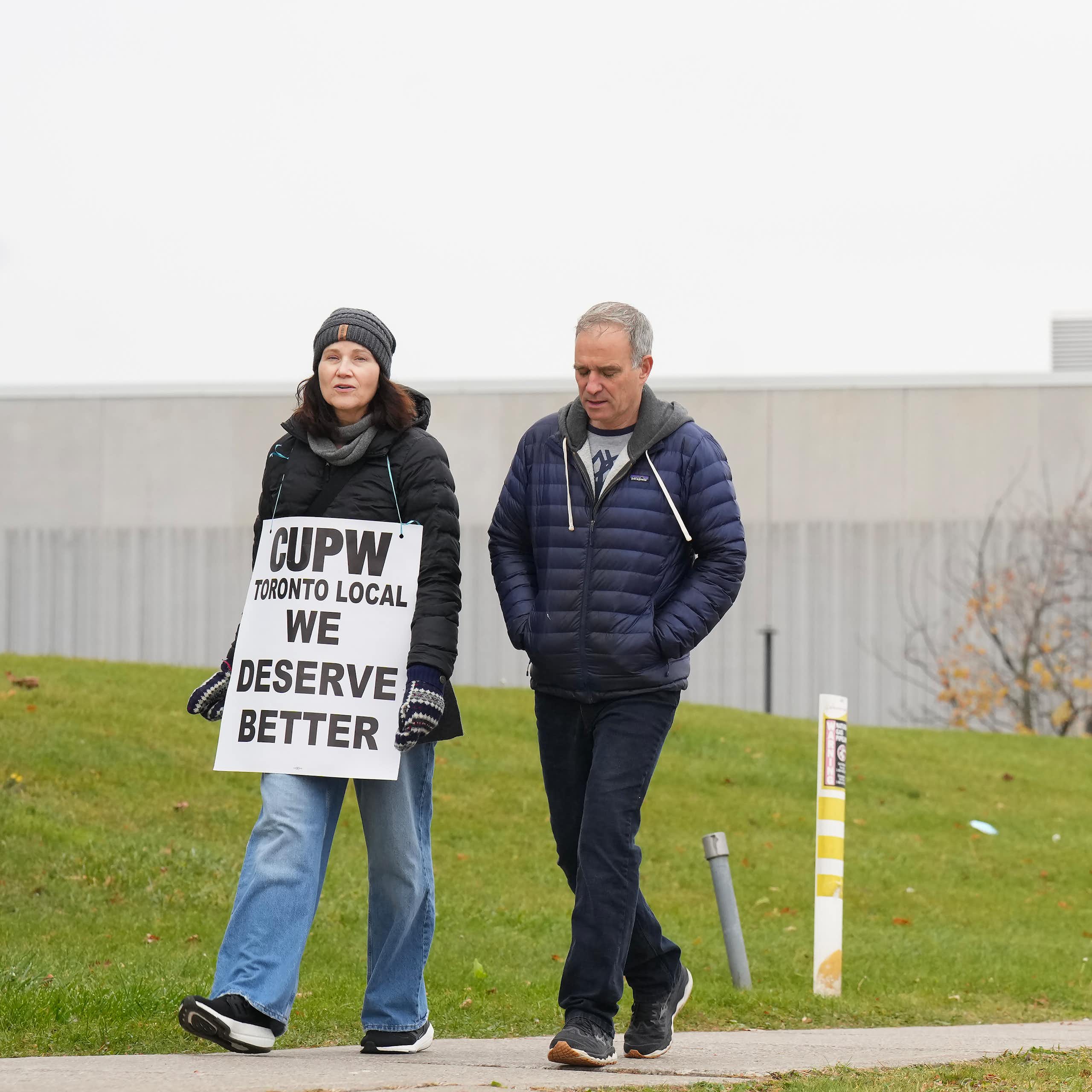 Two people walk on a sidewalk past a Canada Post sign. One of the people is wearing a sign around the neck that says 'CUPW Toronto Local We Deserve Better['