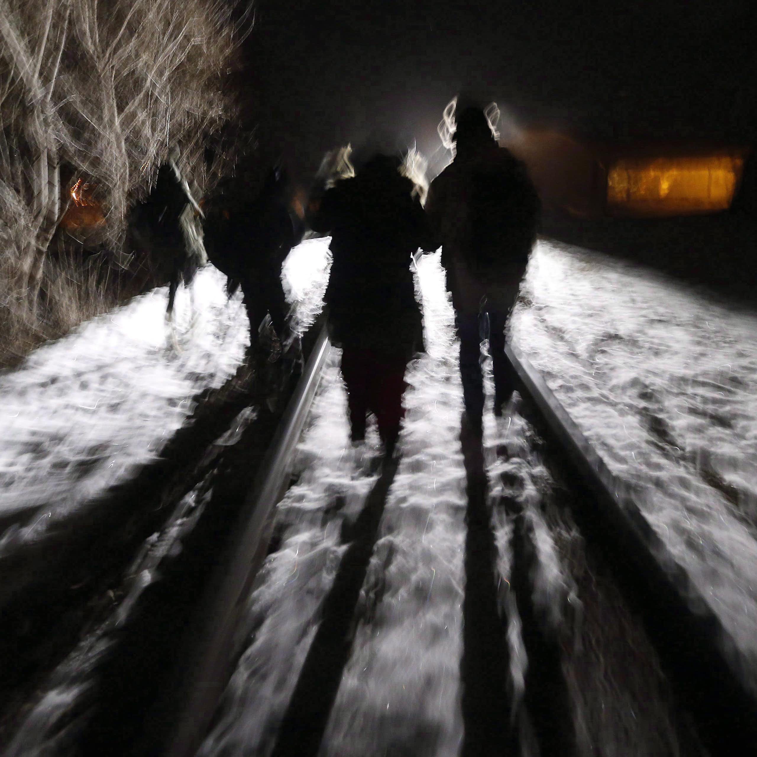 Migrants in silhouette are seen walking on a snowy night along a train tracks. 