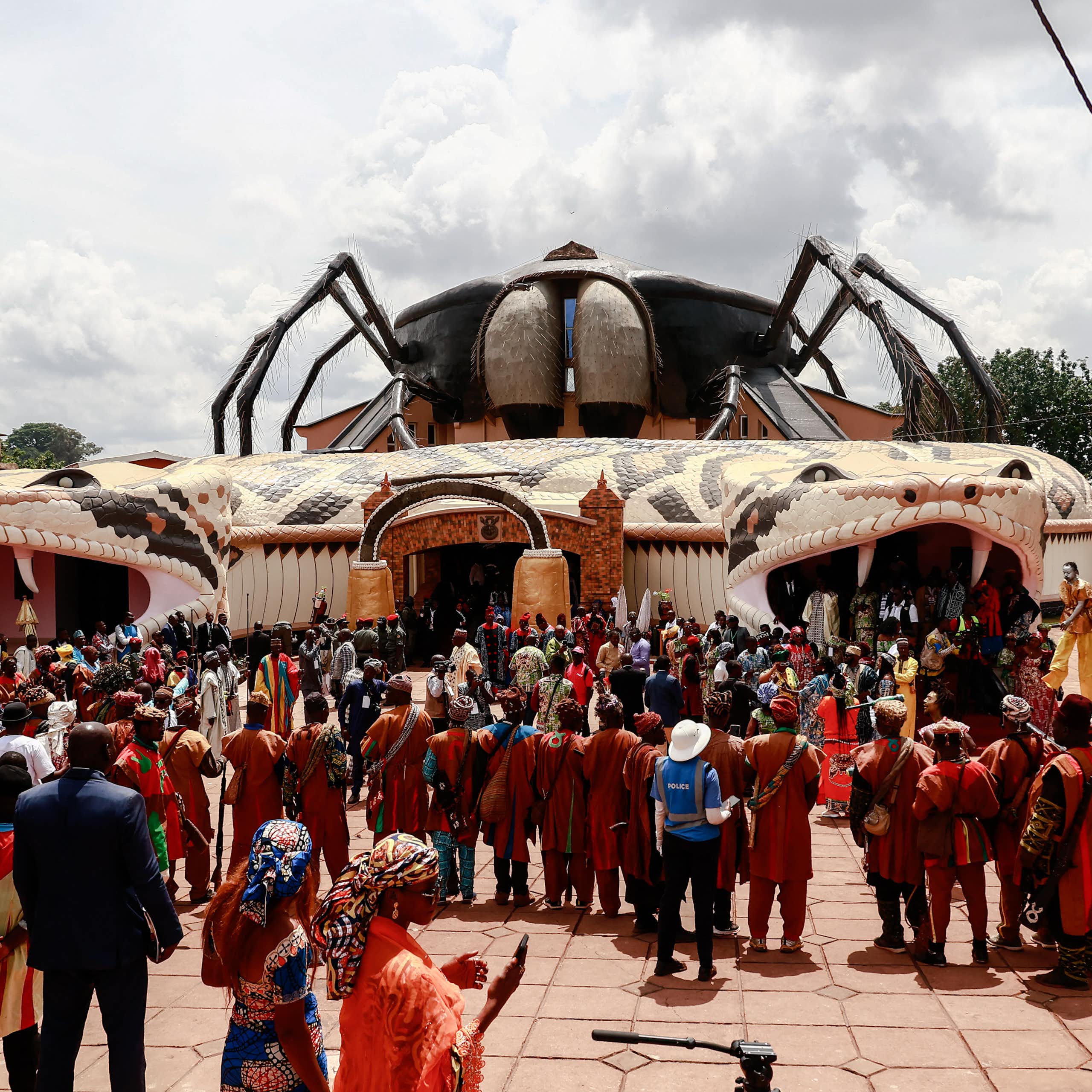 A crowd of people stands outside a curious building, with an entrance and exit on either side through a giant snake's head with fangs that wraps around the building with a huge hairy spider perched on the roof.