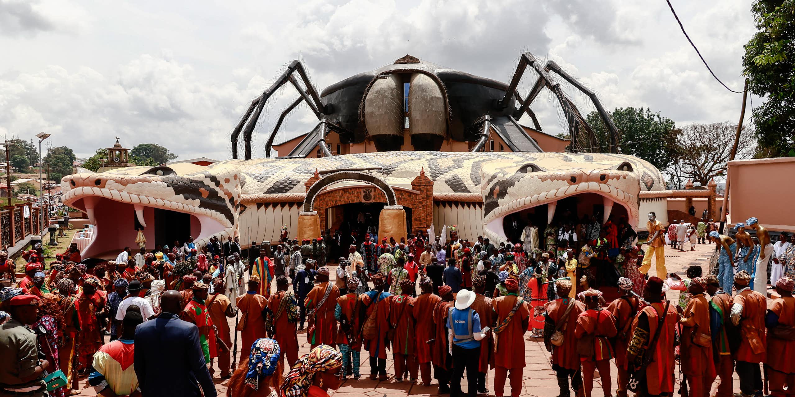 A crowd of people stands outside a curious building, with an entrance and exit on either side through a giant snake's head with fangs that wraps around the building with a huge hairy spider perched on the roof.