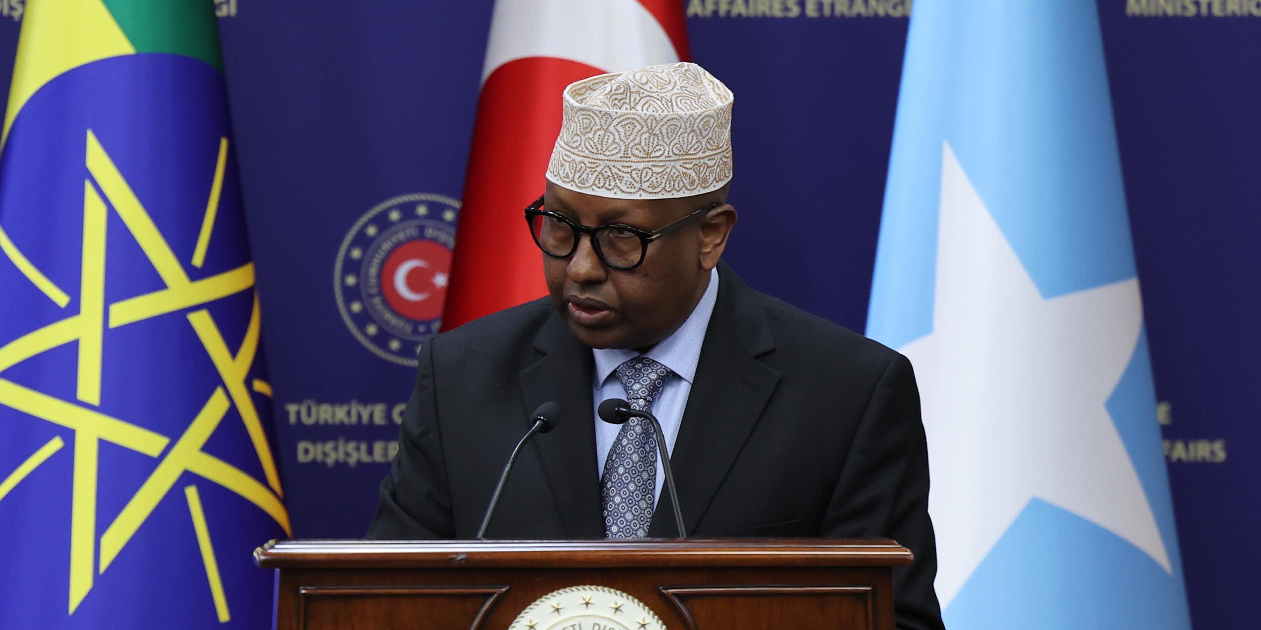 A man speaking into a microphone with three flags behind him