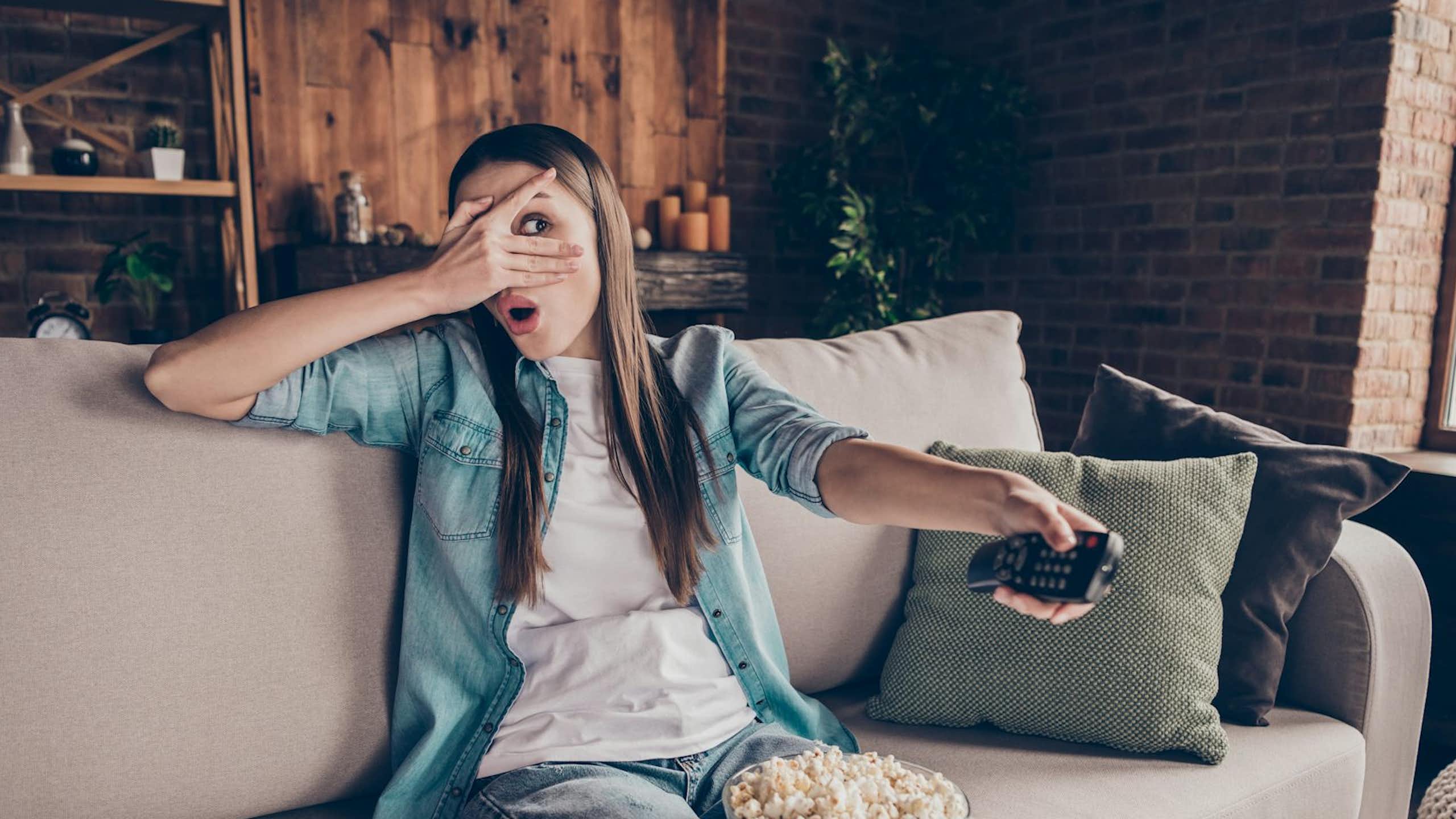 A young woman watches TV with a bowl of popcorn while holding a remote control.