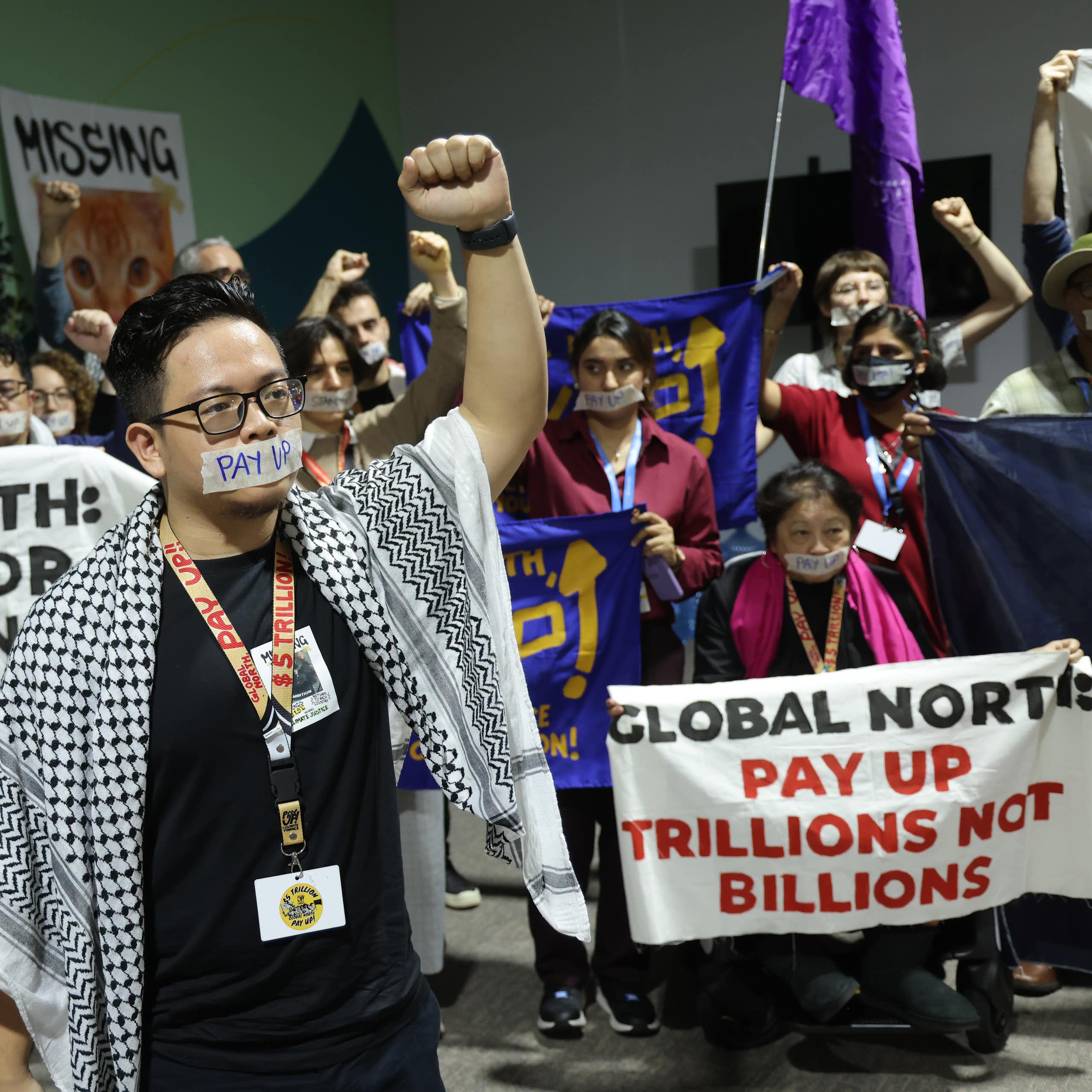 Protestors holding up banners saying 'Global North: pay up trillions not billions' and "Global South: Stand Up for public finance". The protest leader is wearing the Palestinian keffiyeh and their mouth has tape over it.