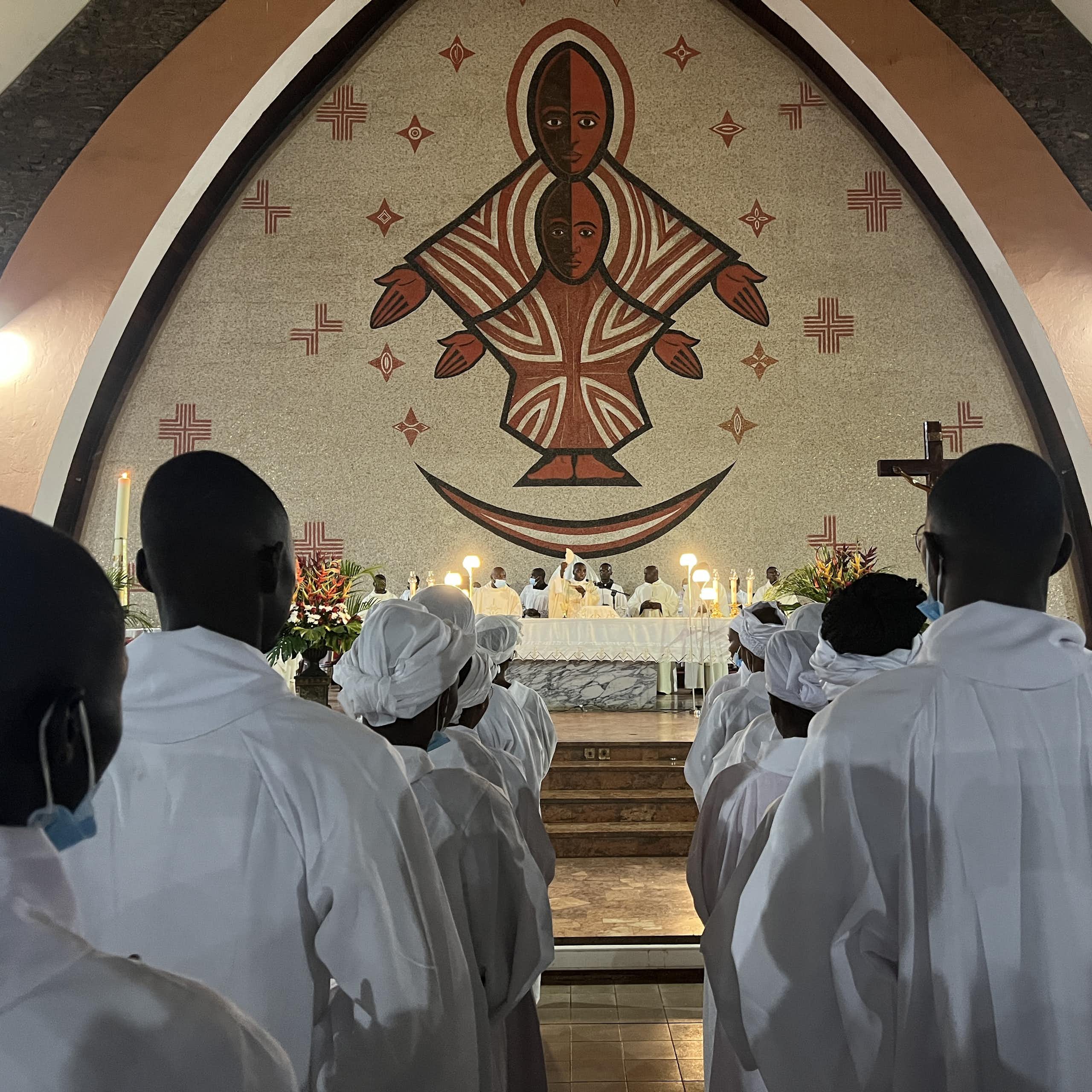 From behind, men and women in white robes line up in a church with candles, a large figurative mural of a black Christ on the wall in front of them.