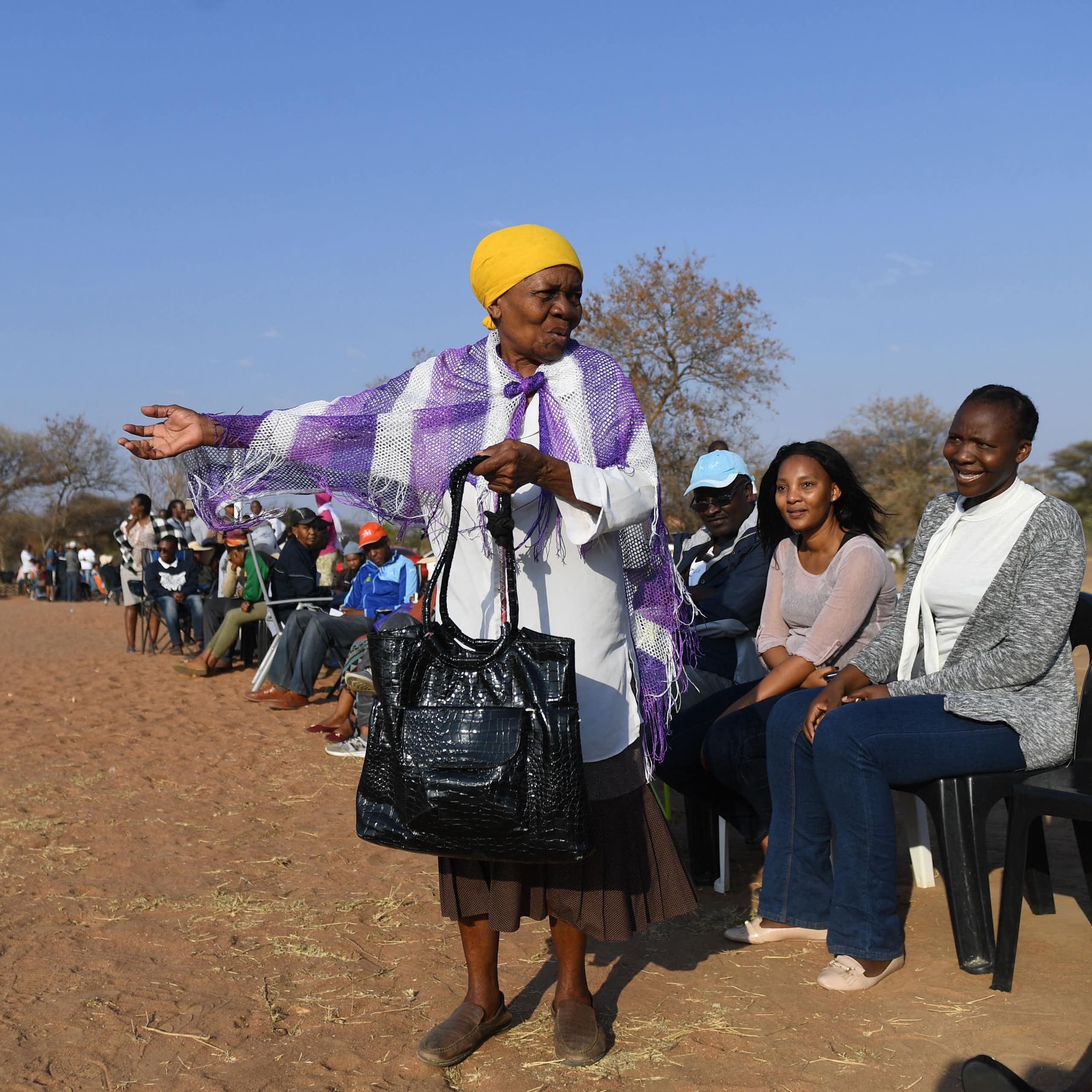 A long queue of seated people on a dusty field. An elderly woman stands in the foreground gesturing and talking.