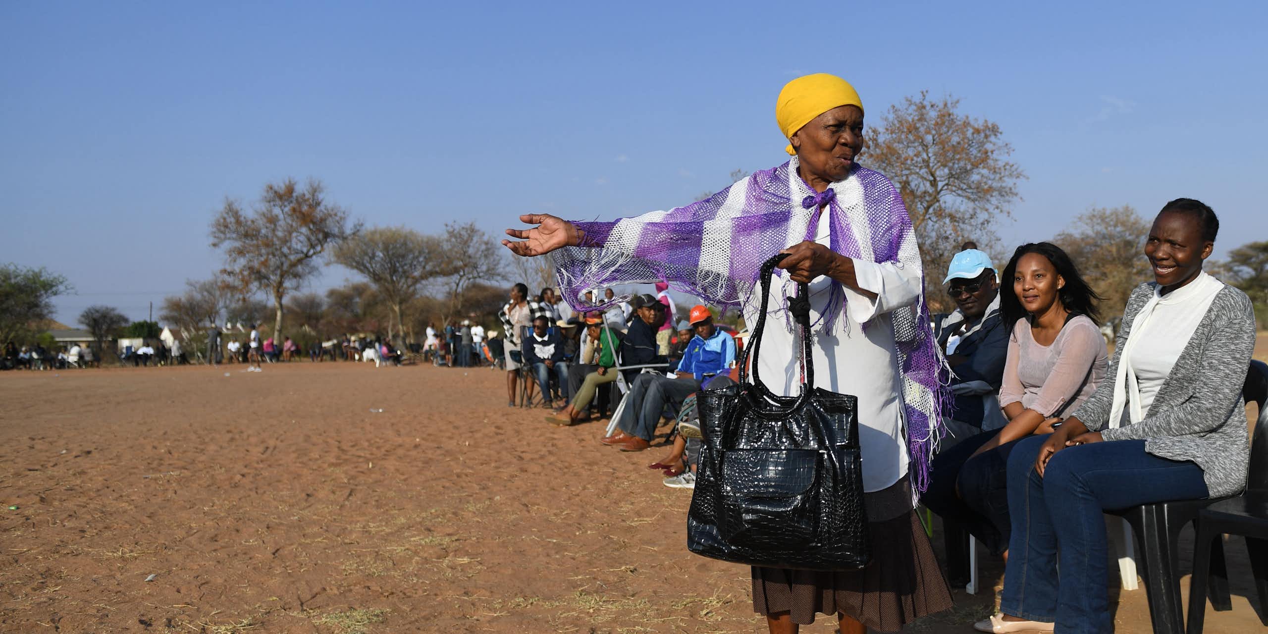 A long queue of seated people on a dusty field. An elderly woman stands in the foreground gesturing and talking.