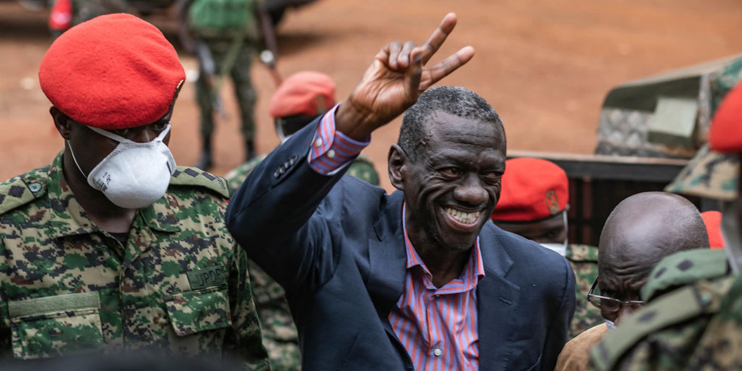 A smiling man holds up two fingers in a wave while surrounded by military men in red caps and military fatigues