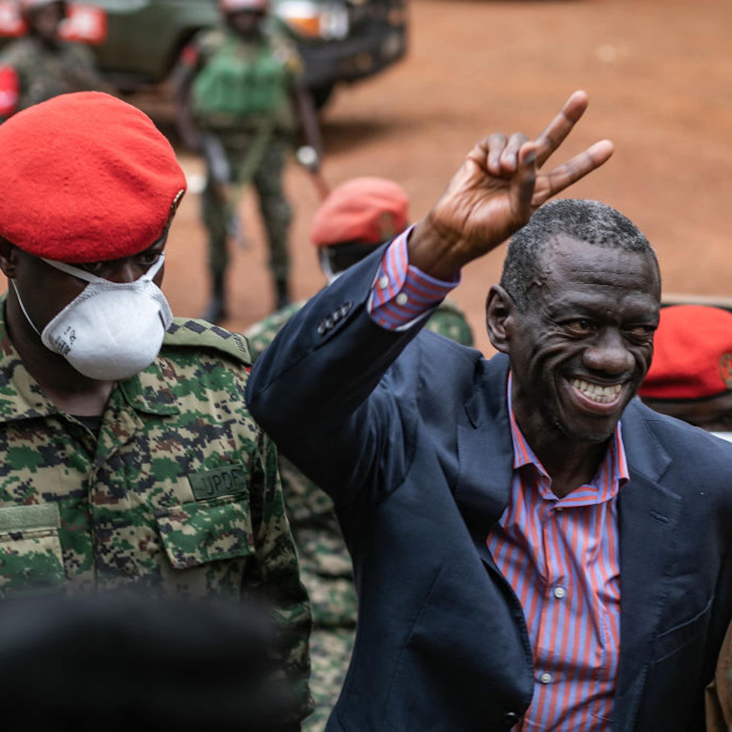 A smiling man holds up two fingers in a wave while surrounded by military men in red caps and military fatigues