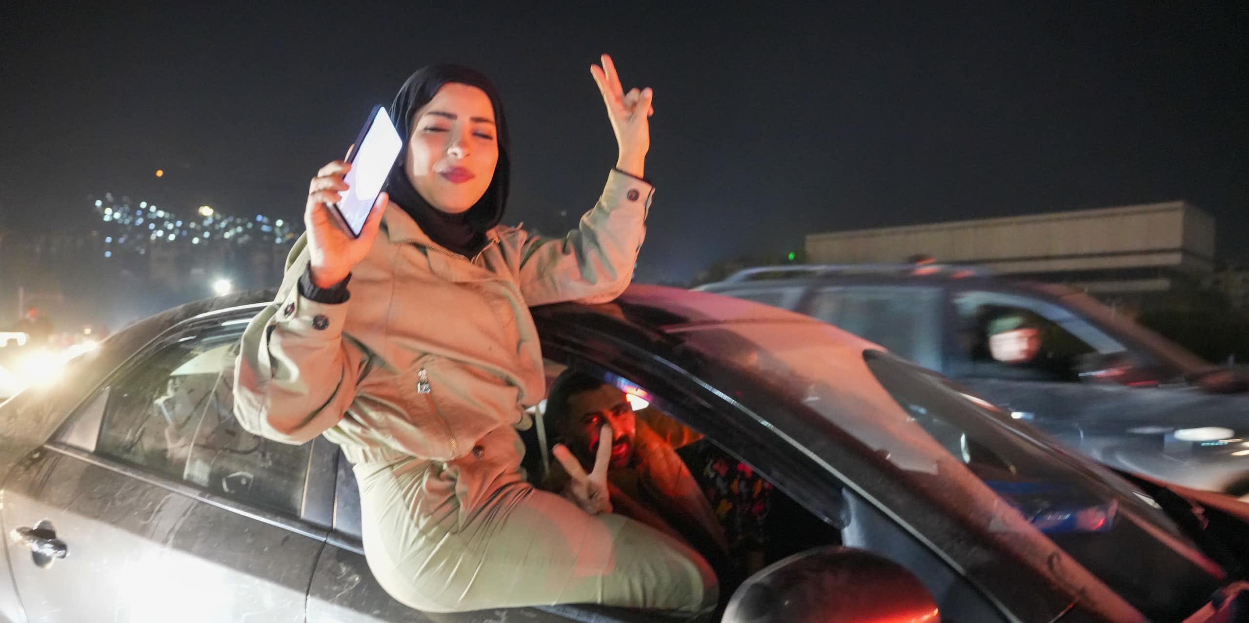 A woman in a tan outfit raising her fingers in a victory sign as she sits in a car window.
