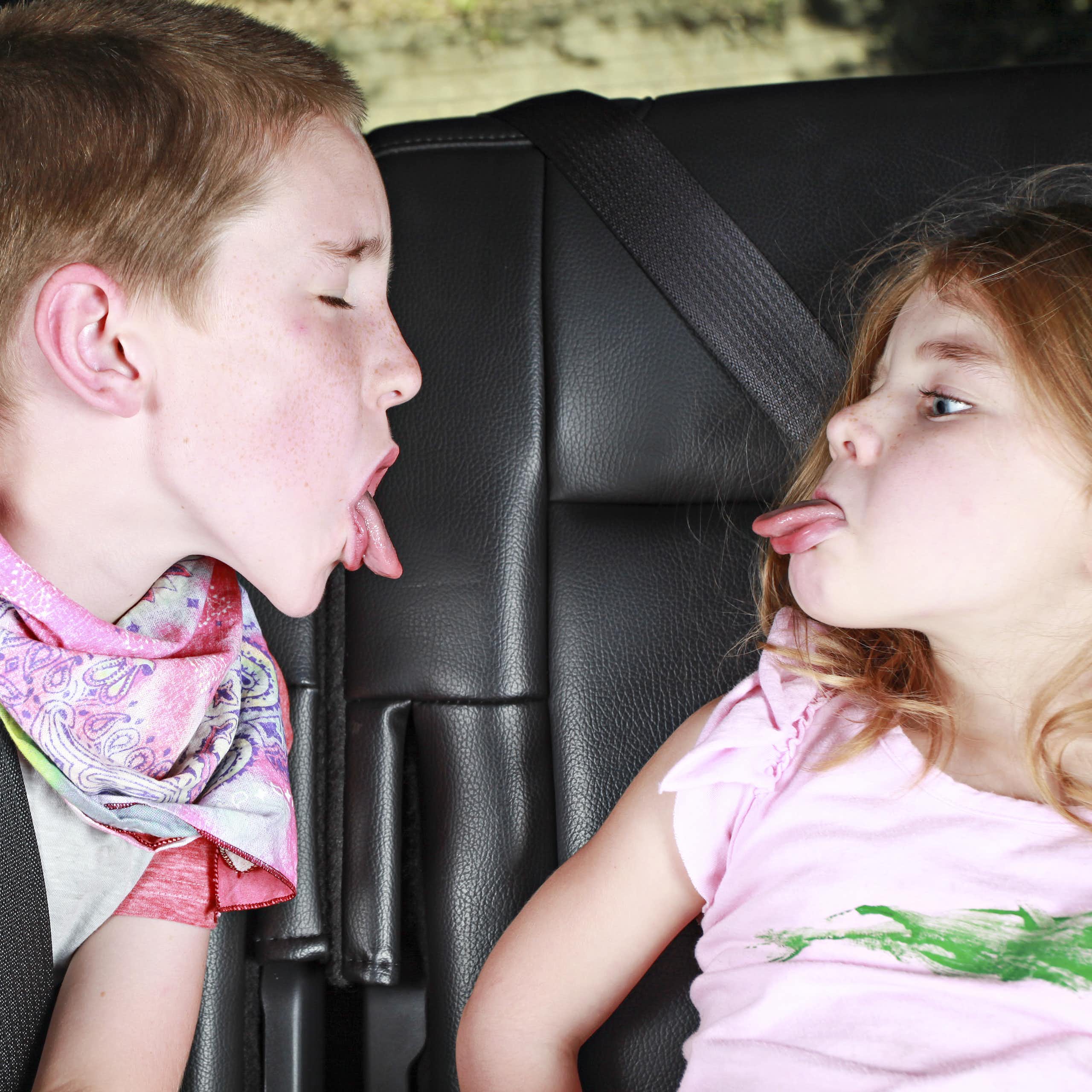 A brother and sister poke their tongues at each other in backseat of a car. 