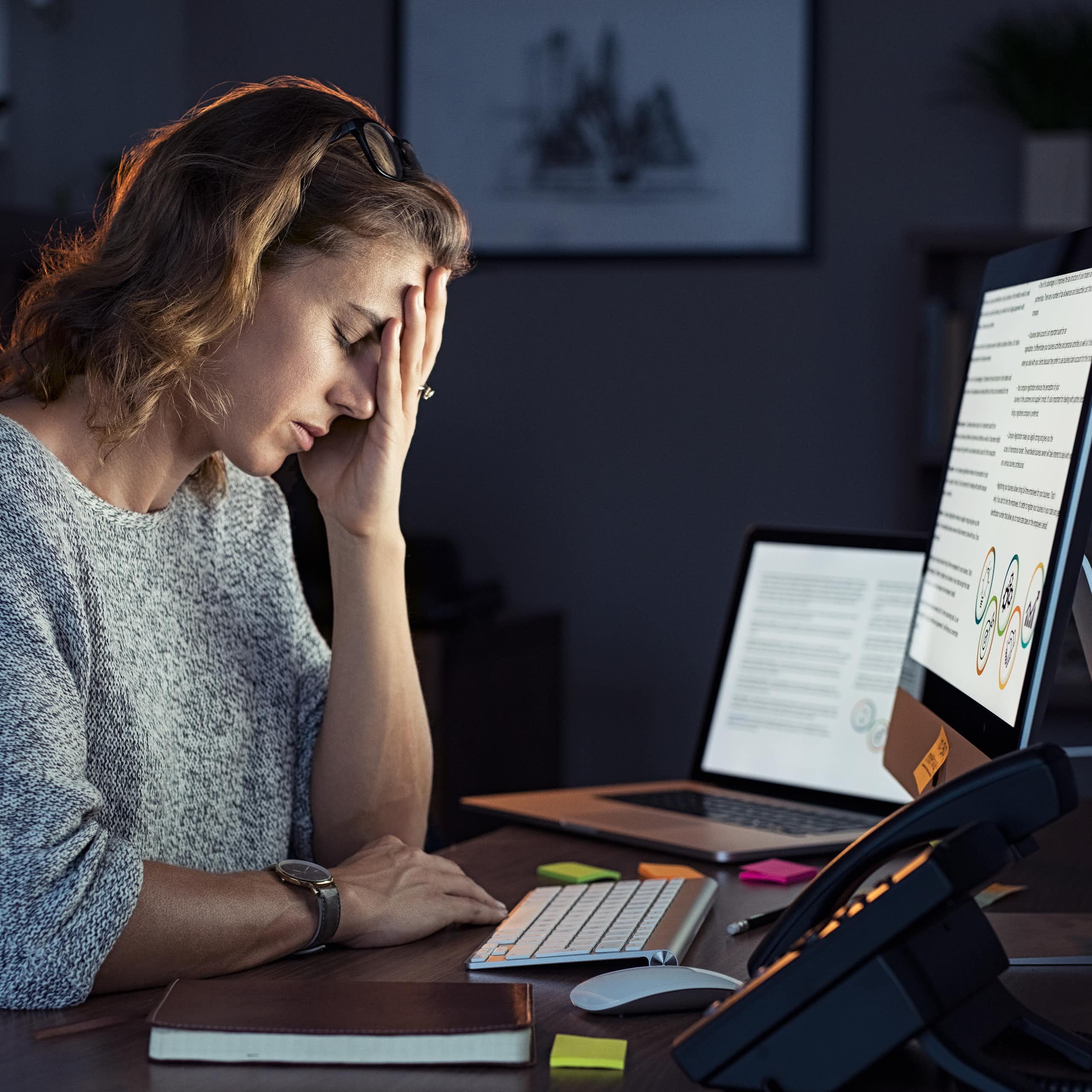 A woman sitting in front of a computer at a desk rests her head against her hand