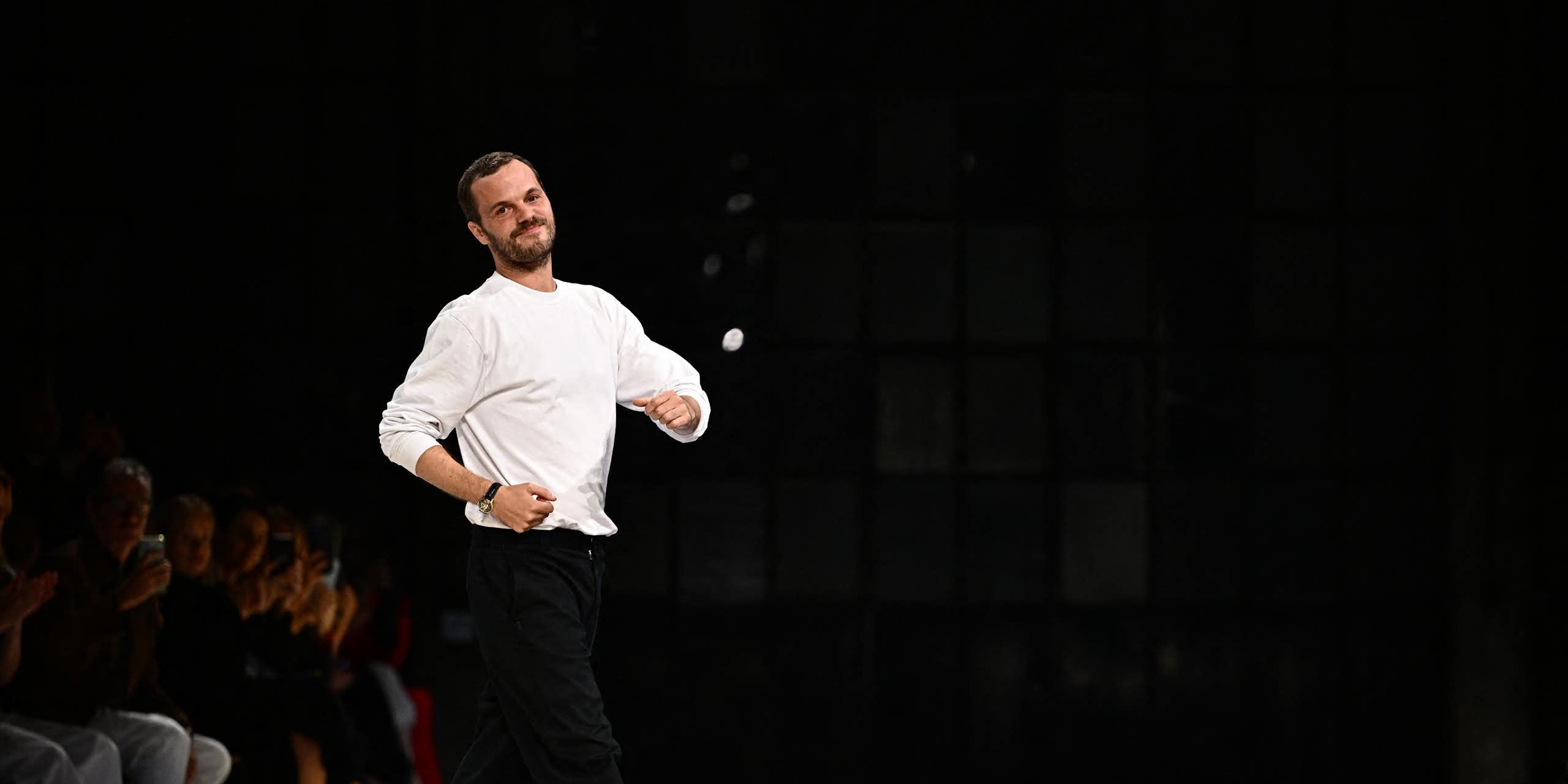 Mathieu Blazy at the end of a runway show, dressed in a white t-shirt and black pants, striking a playful pose.