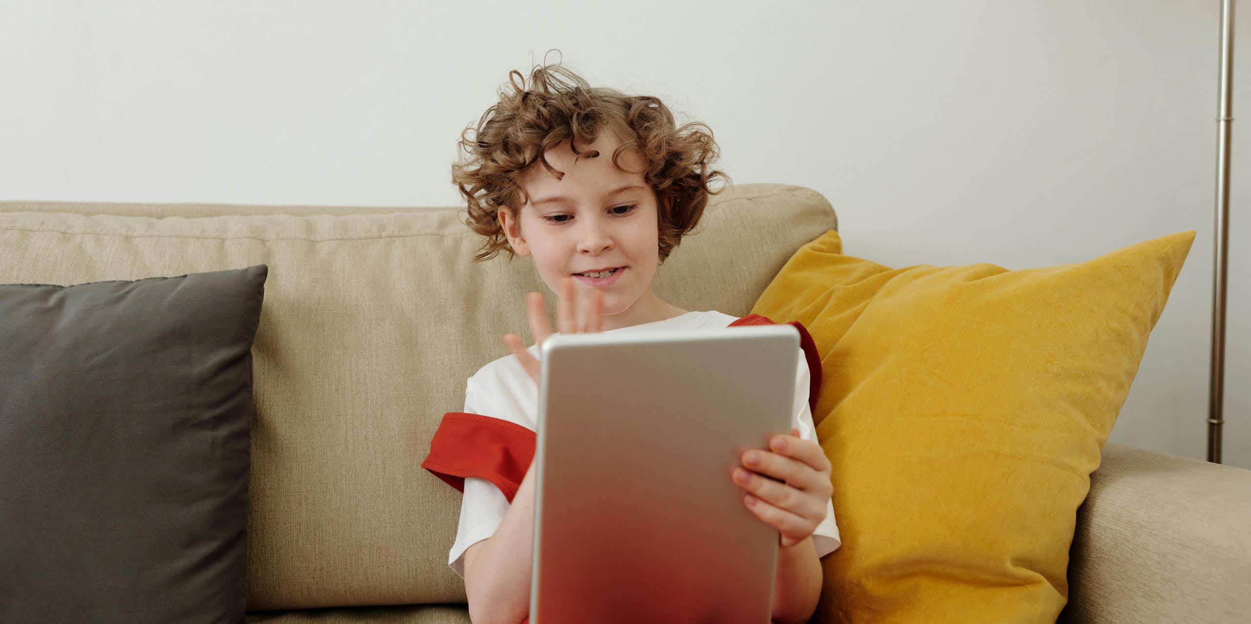 A young child sits on a couch, holding up a tablet and waving. 