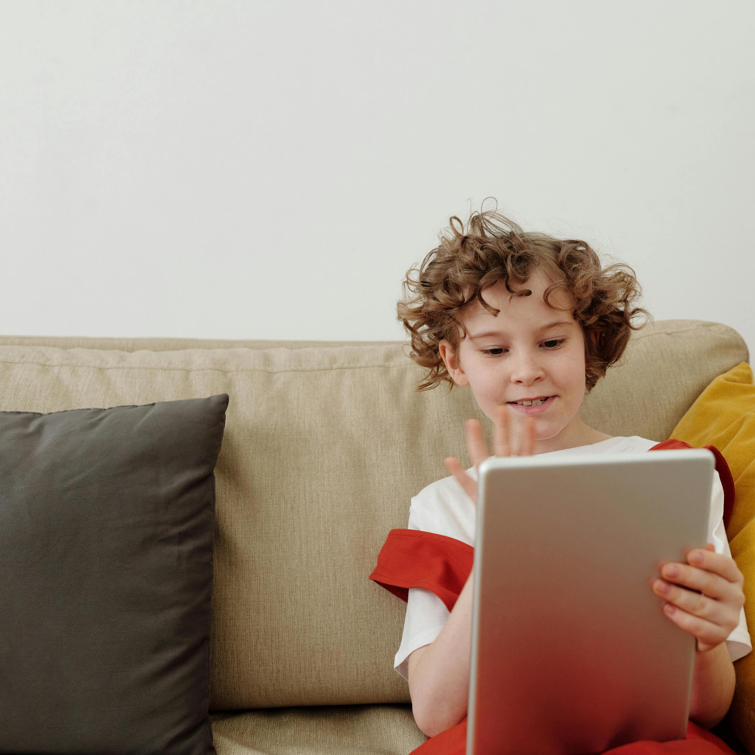 A young child sits on a couch, holding up a tablet and waving. 