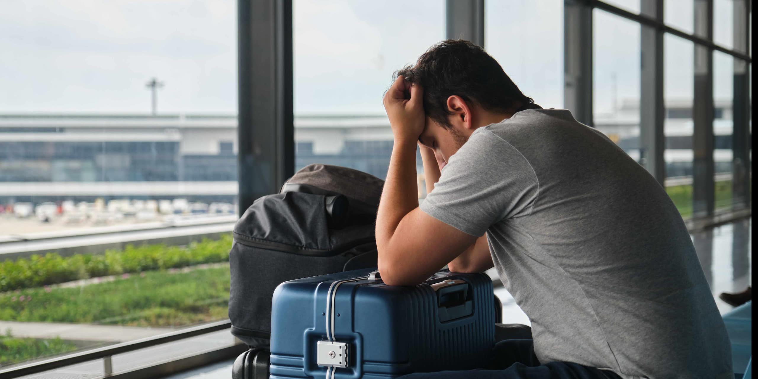 A man in an airport holds his head on top of his suitcase
