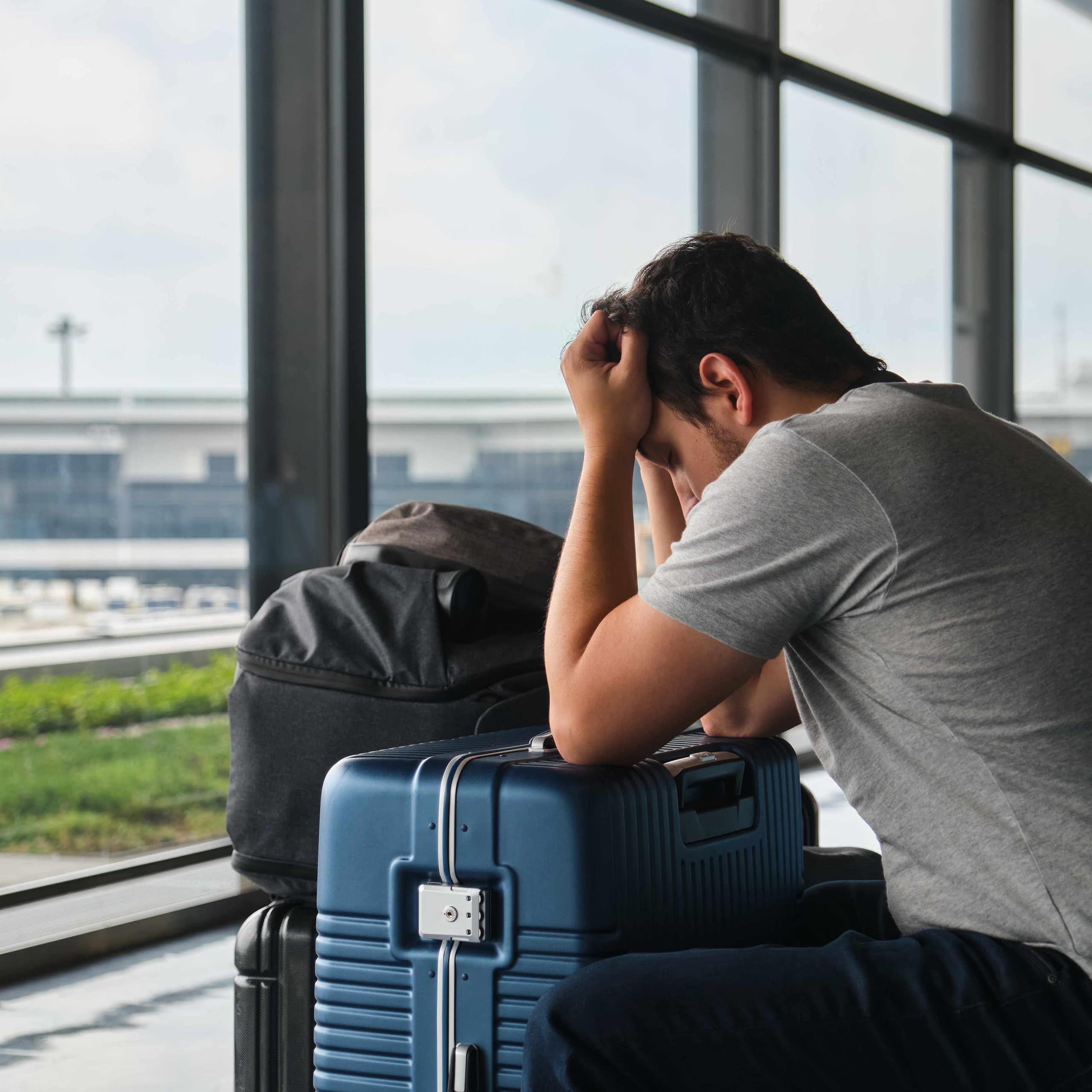A man in an airport holds his head on top of his suitcase