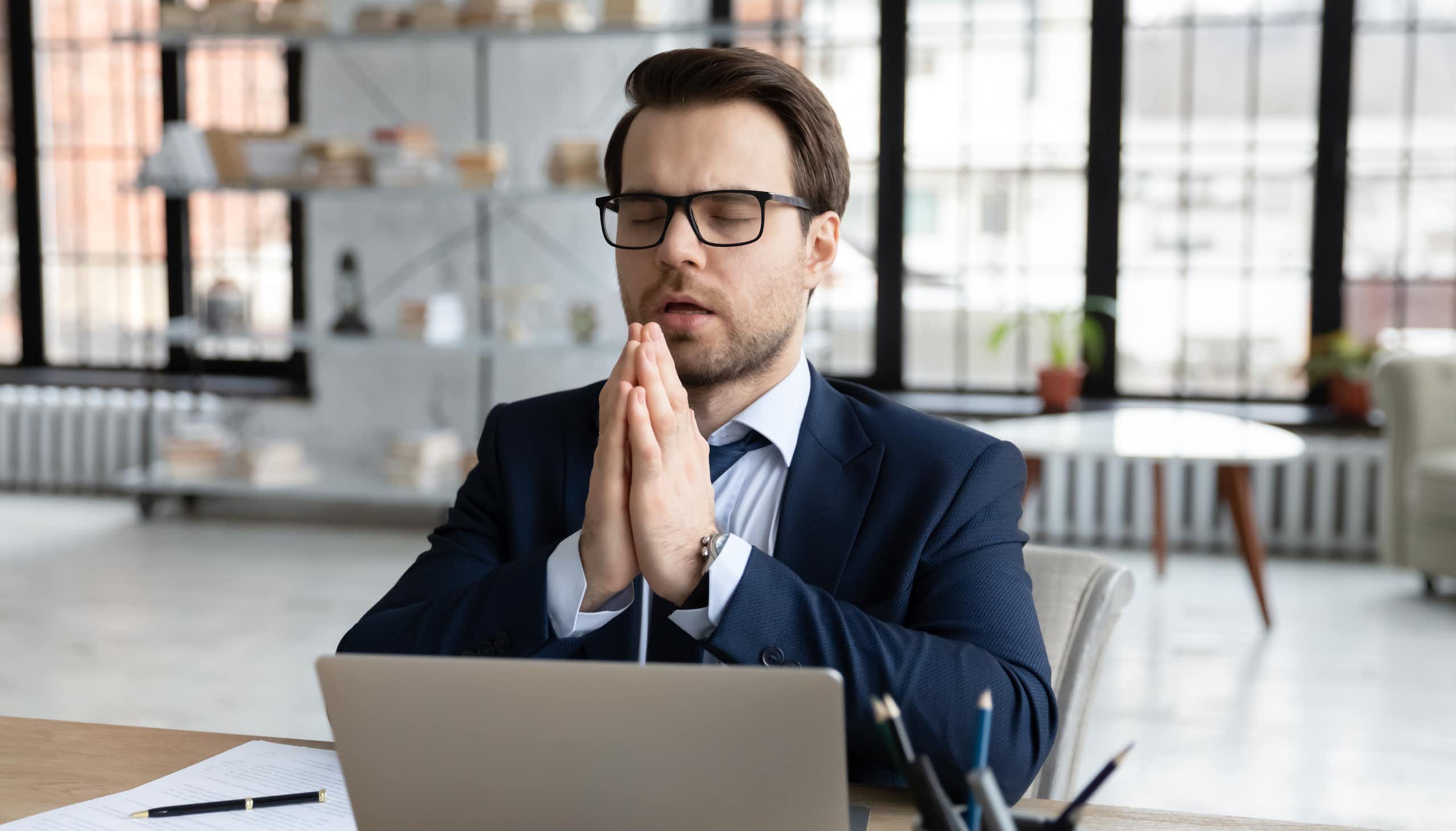 un homme au travail, avec les yeux fermés et les mains jointes dans un environnement de bureaux