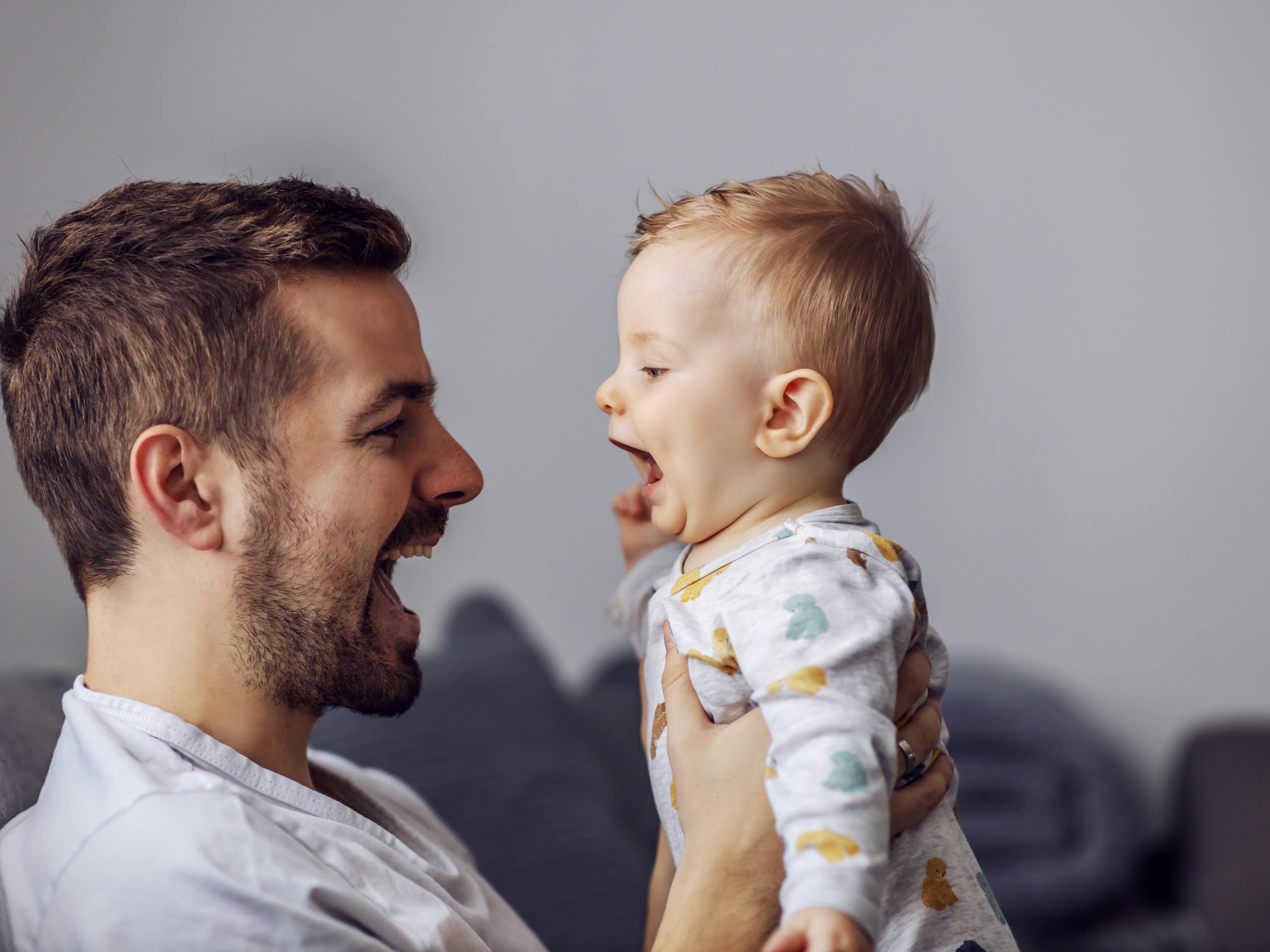 A man holding up a baby, both have their mouths open  talking to each other