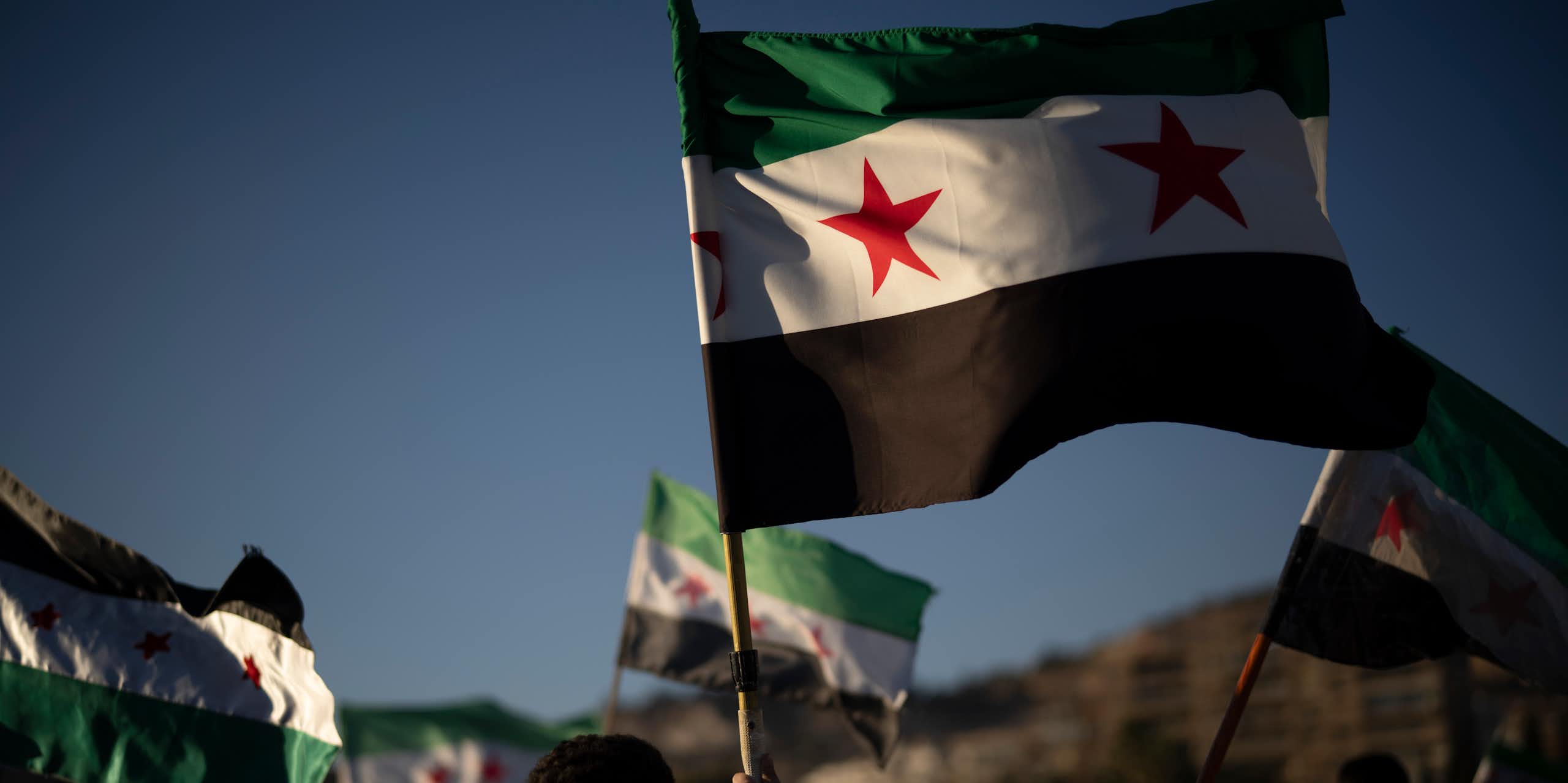 Syrian flags fly against a blue sky, held in the hands of people marching together