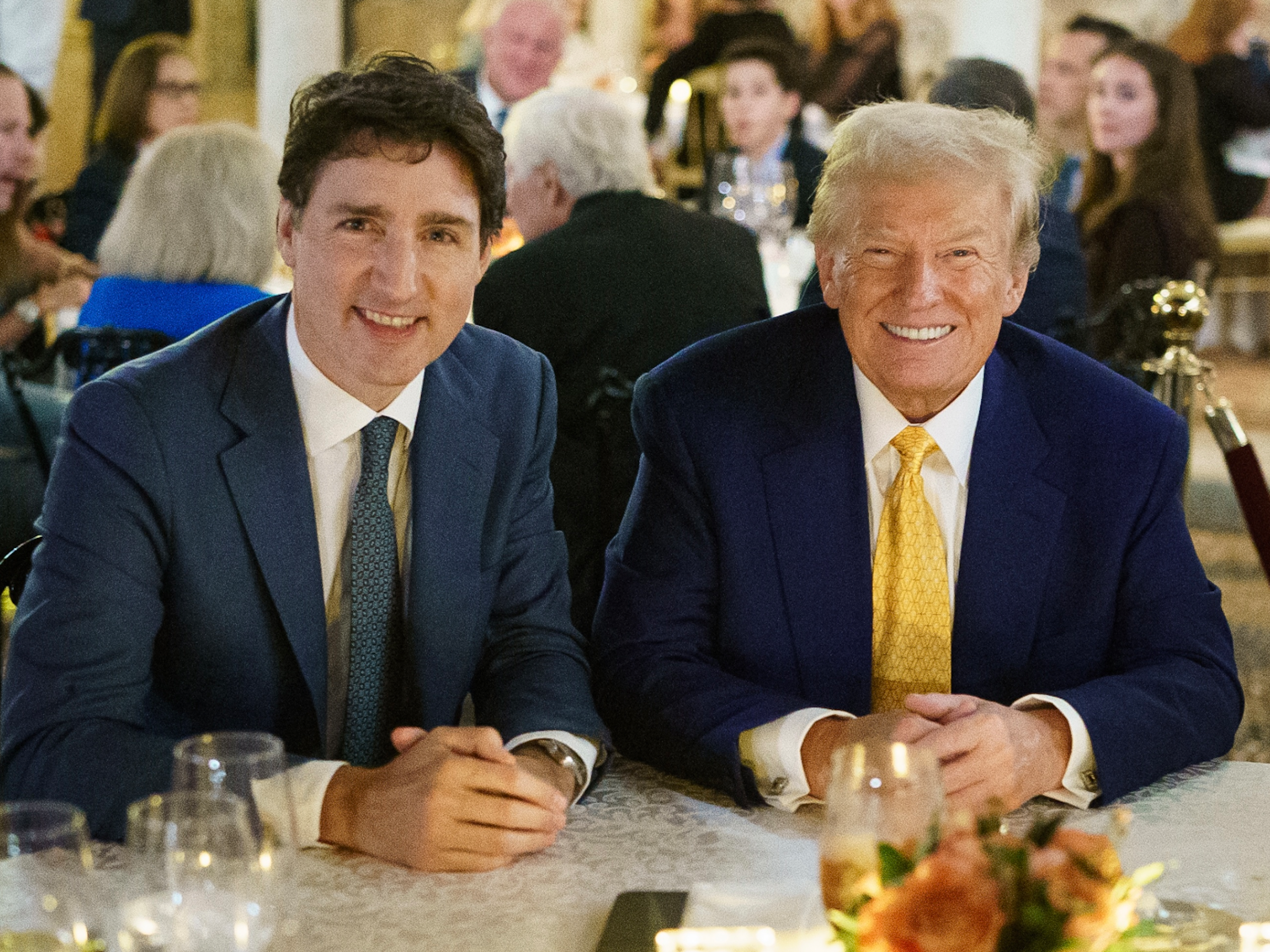 Un homme brun et un homme plus âgé aux cheveux blond-gris sourient à une table.